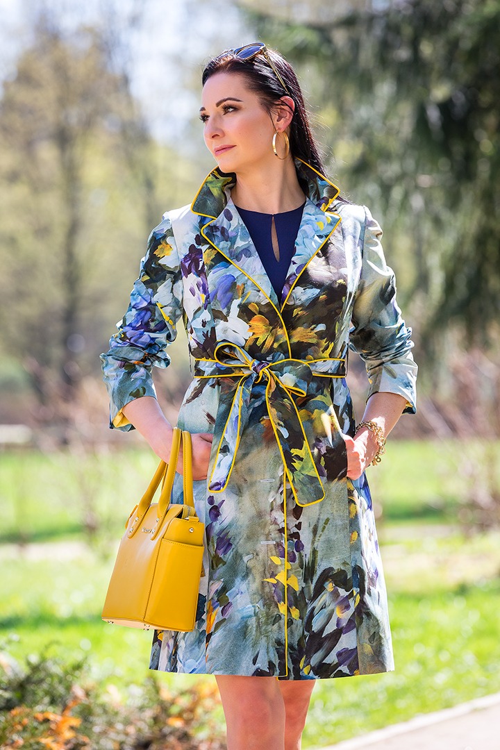 Kobieta w płaszczu w kwiaty z żółtą lamówką i torebką w kolorze musztardowym, na tle parku.