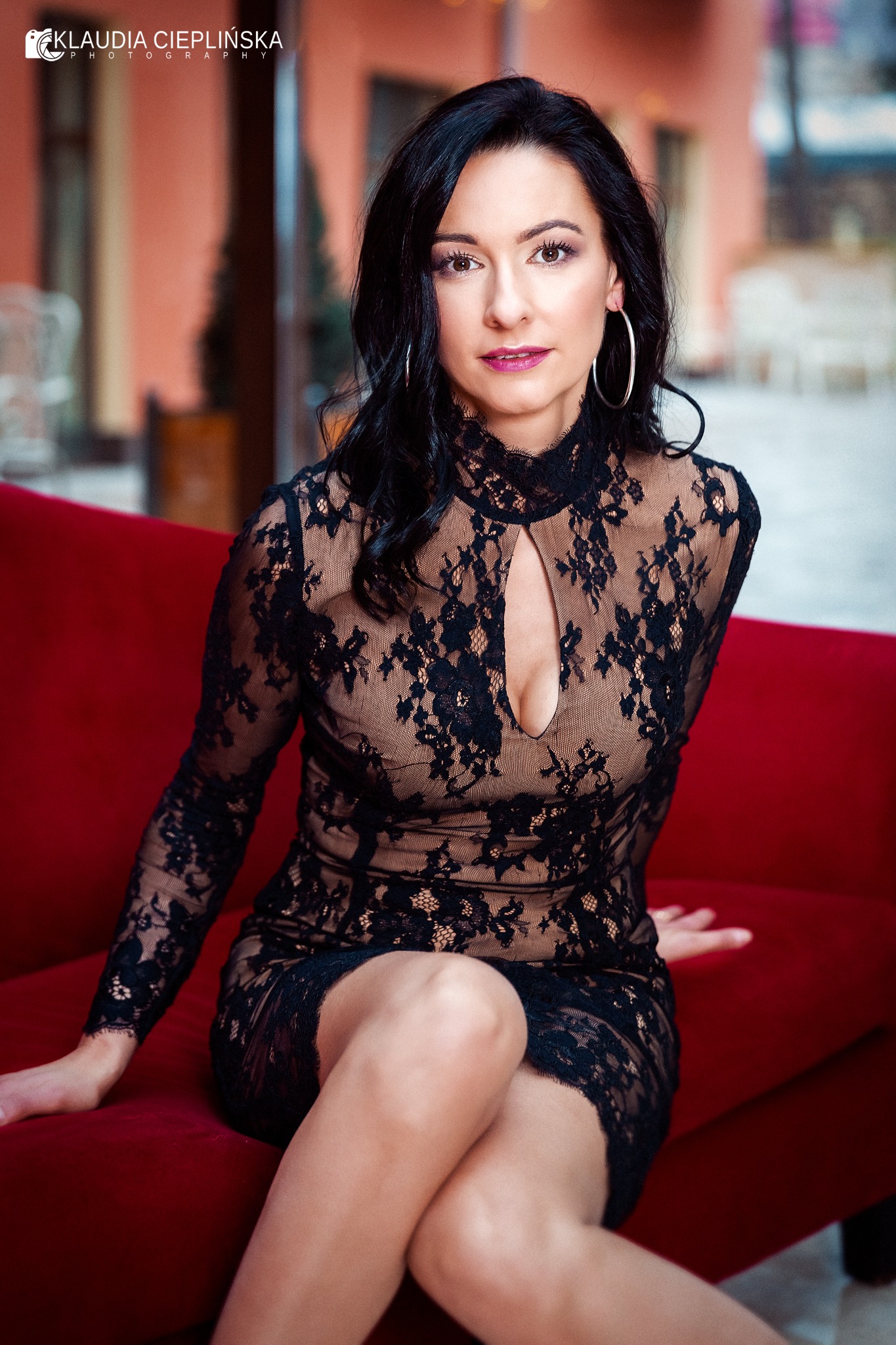 Kobieta w czarnej koronkowej sukience z długim rękawem i wycięciem na dekolcie, siedząca na czerwonej sofie, z dużymi srebrnymi kolczykami kołami.