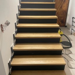 FrenchTouch - Nowoczesne schody z drewnianymi stopniami i czarnymi podstopnicami, widok z dołu, z widoczną instalacją elektryczną i drzwiami w tle.