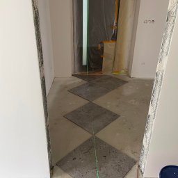 FrenchTouch - Układanie szarych, kwadratowych płytek podłogowych w romby, wyznaczane laserem krzyżowym. Drzwi zabezpieczone folią malarską. Widoczny fragment puszki z farbą.