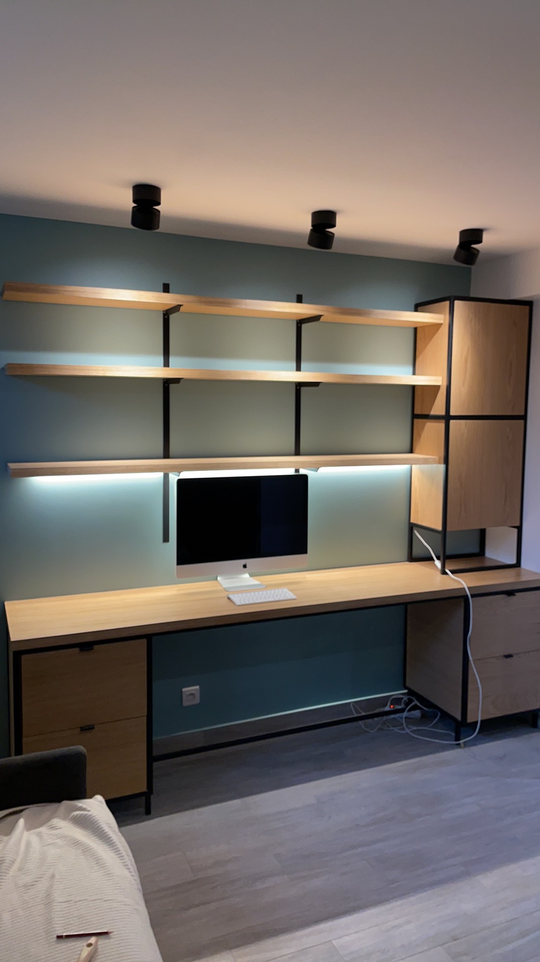 Nowoczesne biuro z drewnianym biurkiem, półkami i szafką w czarnych ramach. Oświetlenie punktowe i listwa LED. Na biurku komputer iMac z klawiaturą.