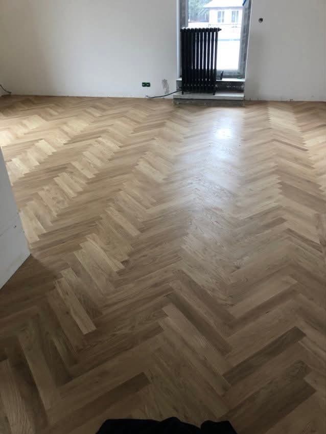 Ułożony parkiet w jodełkę francuską, jasne drewno, widok na grzejnik pod oknem. Minimalistyczne wnętrze po renowacji podłogi.