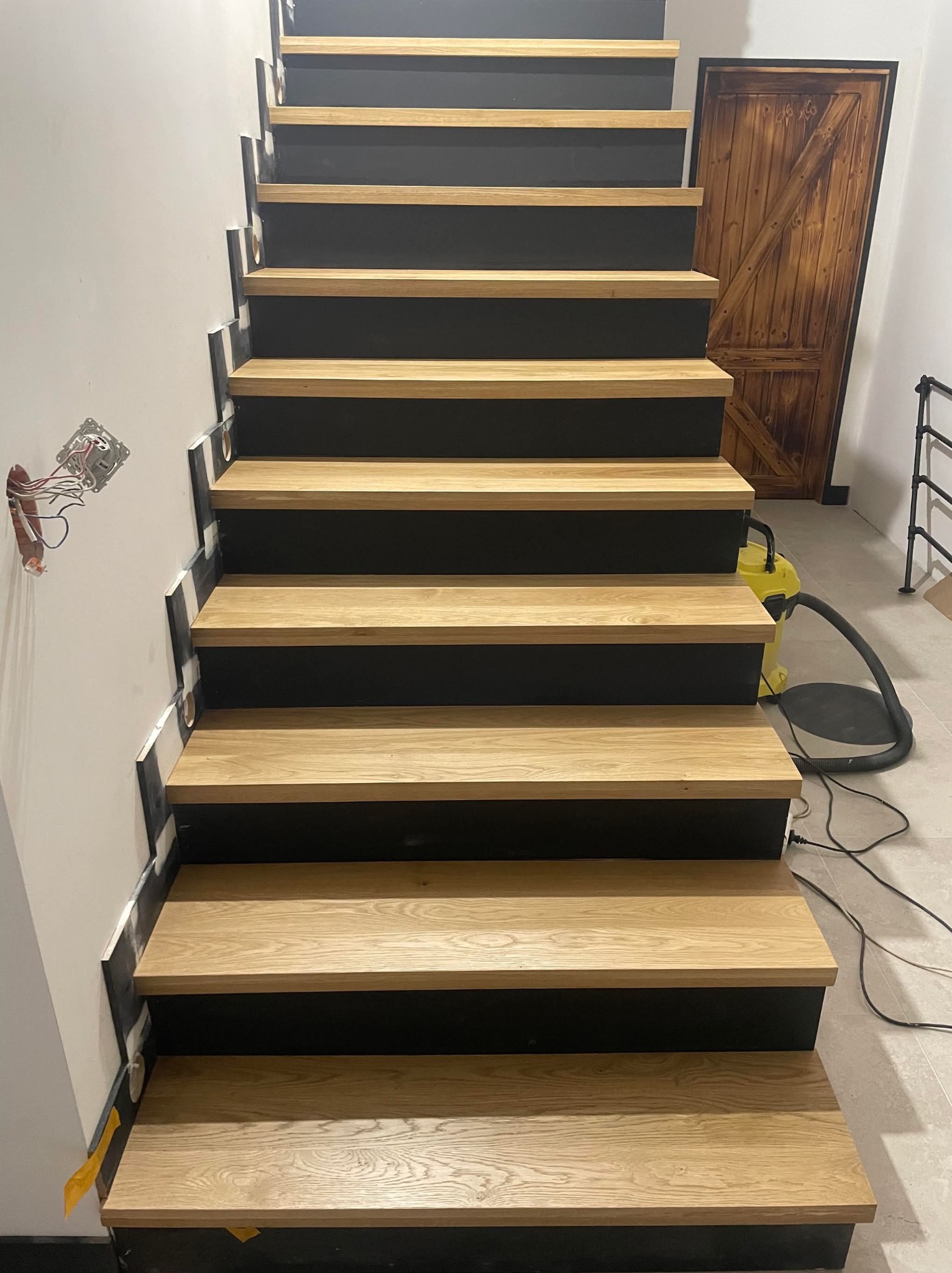 Nowoczesne schody z drewnianymi stopniami i czarnymi podstopnicami, widok z dołu, z widoczną instalacją elektryczną i drzwiami w tle.