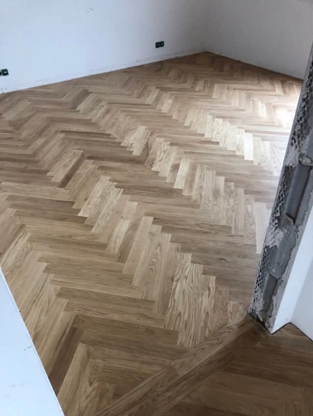 Ułożony parkiet w jodełkę francuską, jasne drewno, widoczne gniazdka elektryczne na ścianie, fragment futryny drzwiowej w tle. Naturalne oświetlenie.