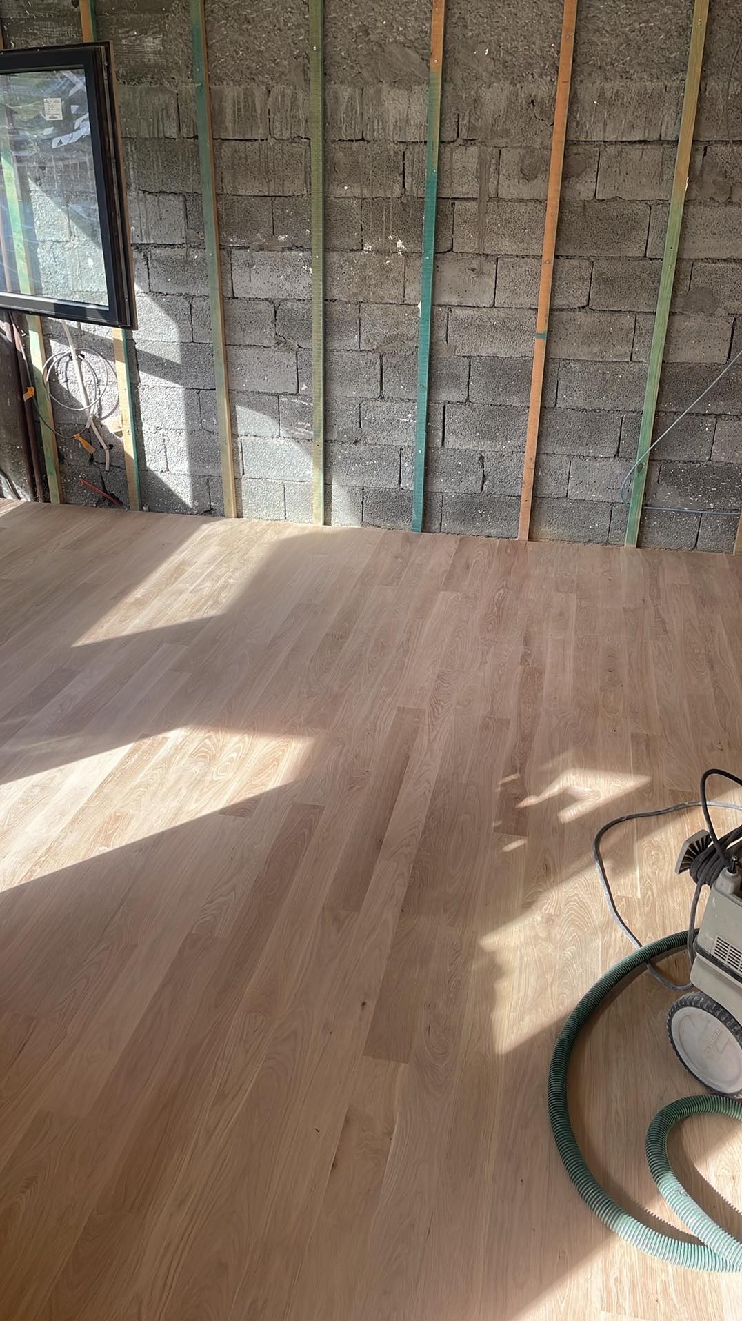 Jasny parkiet ułożony w surowym wnętrzu z betonową ścianą i drewnianymi listwami. W rogu stoi maszyna do polerowania podłóg.
