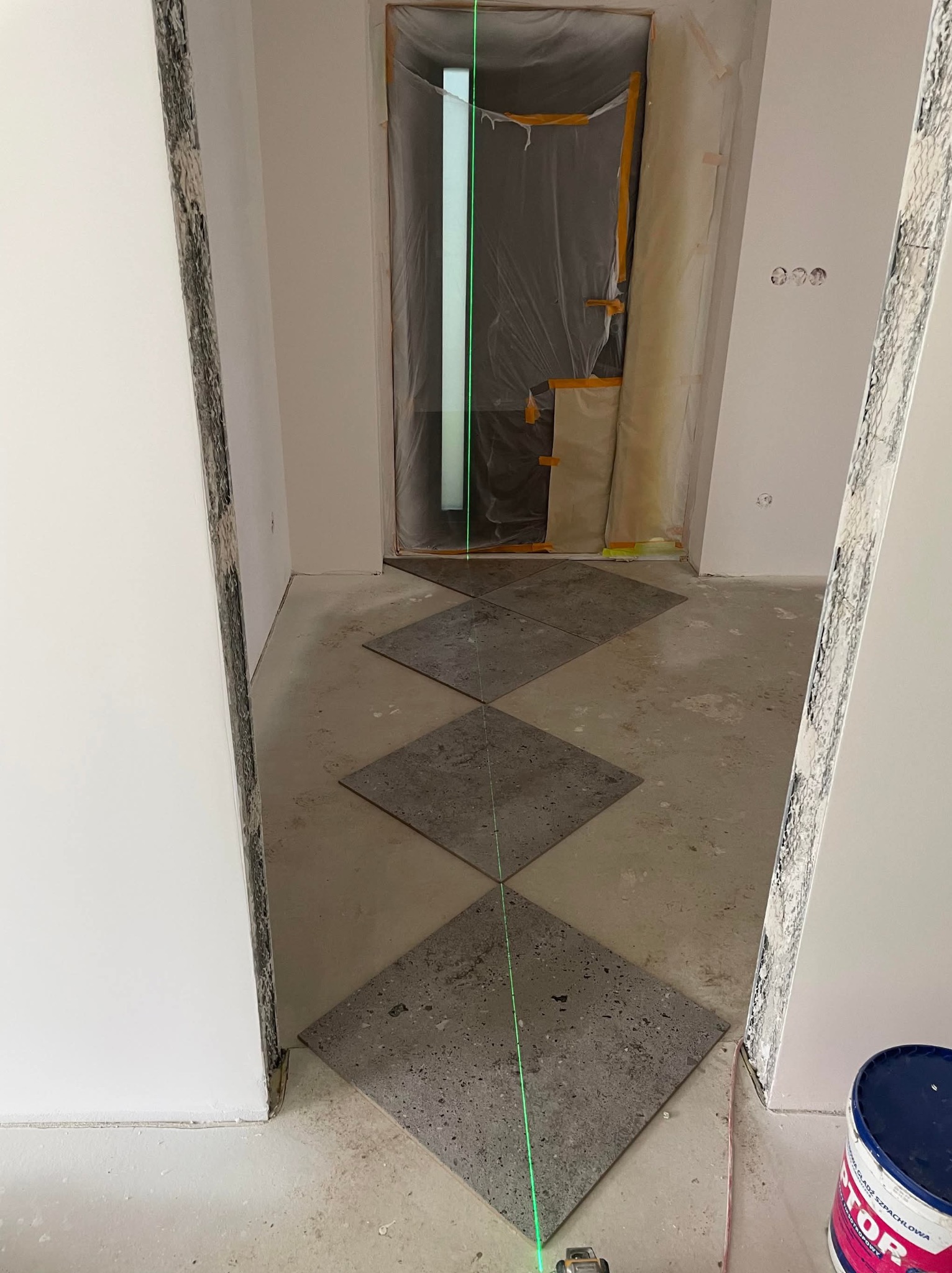 Układanie szarych, kwadratowych płytek podłogowych w romby, wyznaczane laserem krzyżowym. Drzwi zabezpieczone folią malarską. Widoczny fragment puszki z farbą.