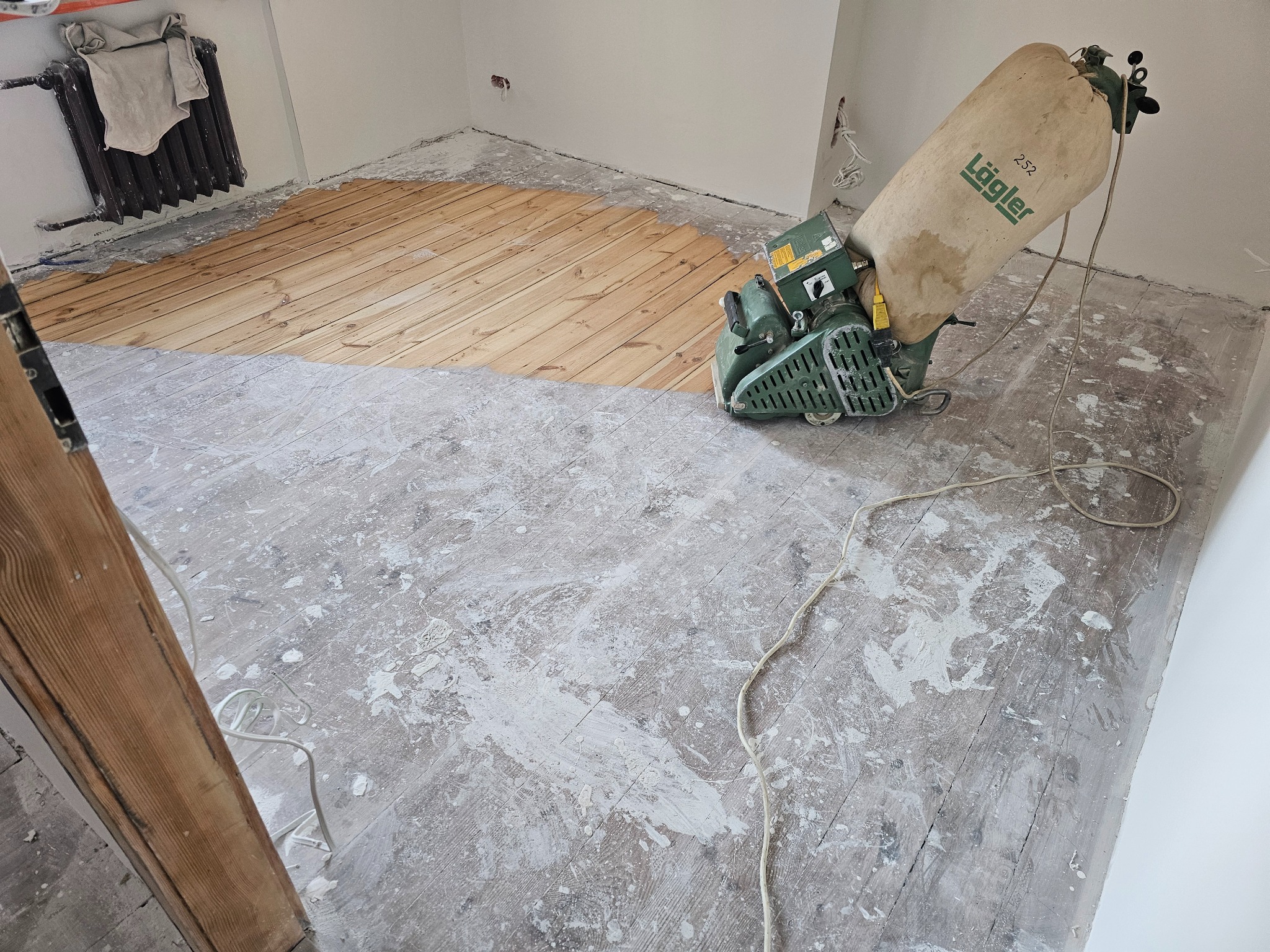 Parkiet w trakcie renowacji: połowa powierzchni wycyklinowana, ukazująca naturalne drewno, druga połowa pokryta resztkami starej farby i pyłu po szlifowaniu. W rogu pokoju stoi grzejnik, a na nim...