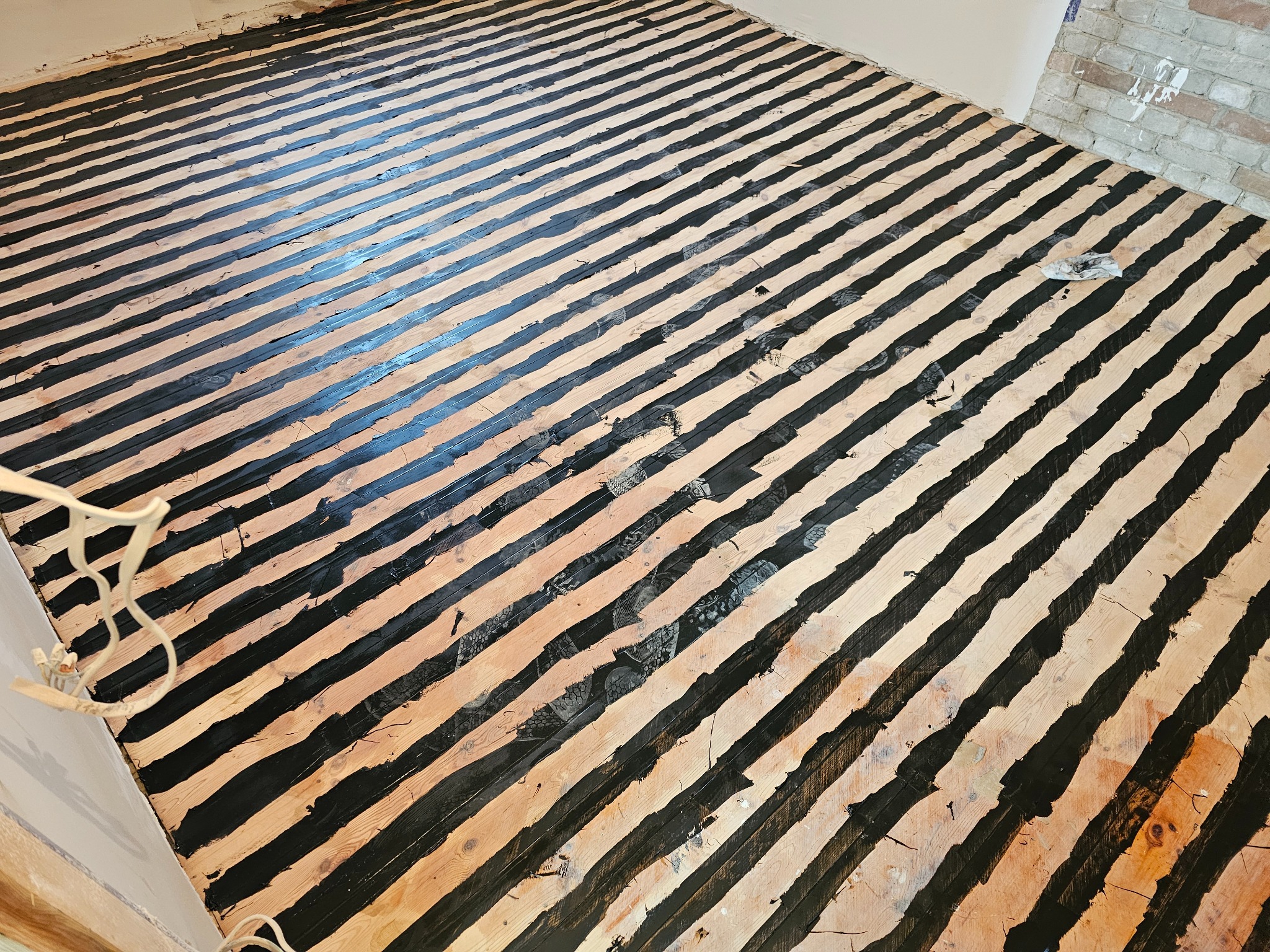 Pomalowane na czarno deski podłogowe układane w pasy, widoczne ślady butów i niedomalowania, fragment ściany z cegły i wystający kabel oświetleniowy.