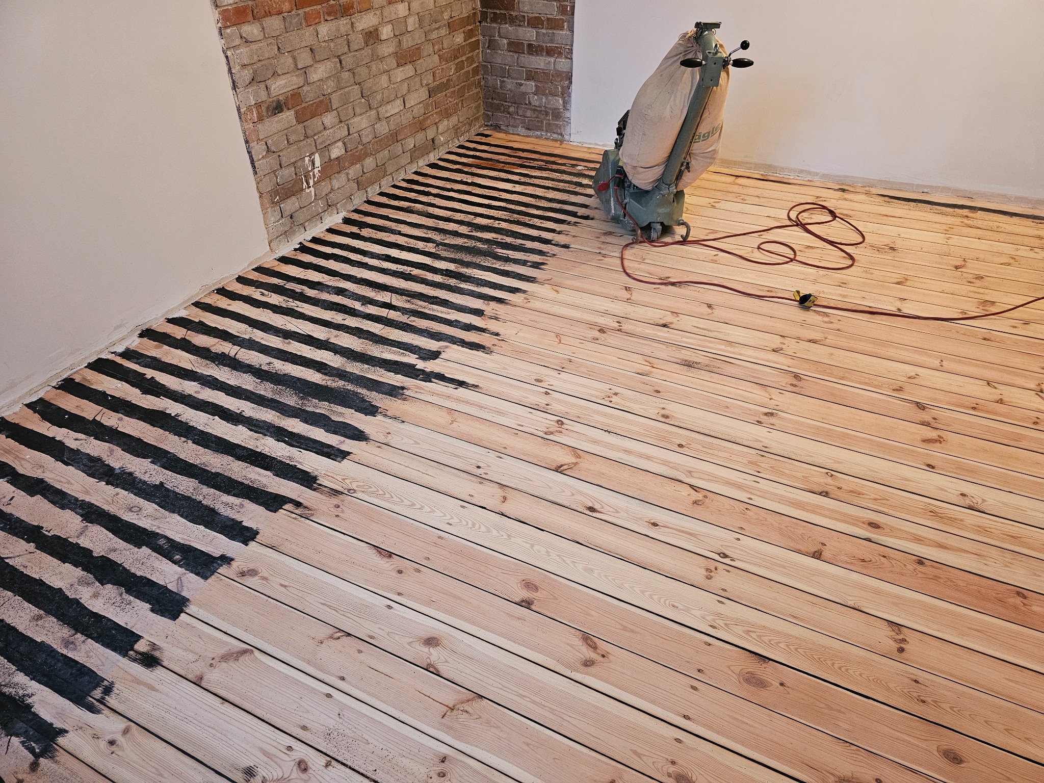 Drewniana podłoga w trakcie renowacji, widoczny fragment po cyklinowaniu i fragment z ciemnymi pasami po wstępnym szlifowaniu, w tle ceglana ściana i maszyna do cyklinowania.