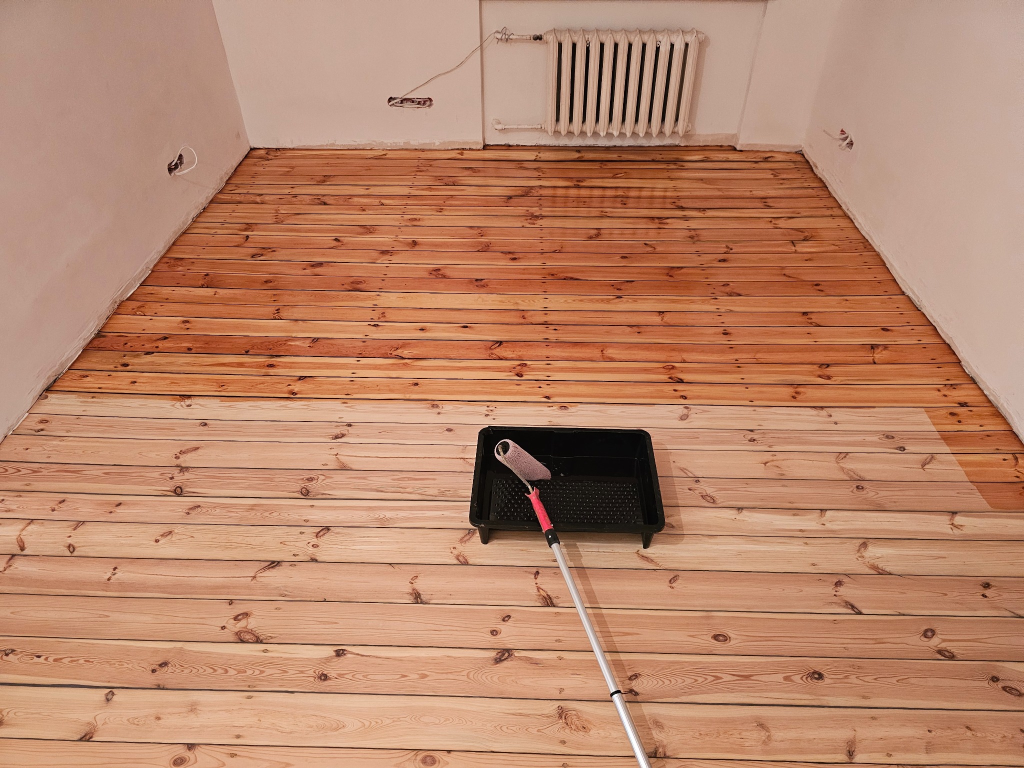 Częściowo polakierowana drewniana podłoga z widocznym wałkiem malarskim w kuwecie, ukazująca różnicę w kolorze między surowym a polakierowanym drewnem.