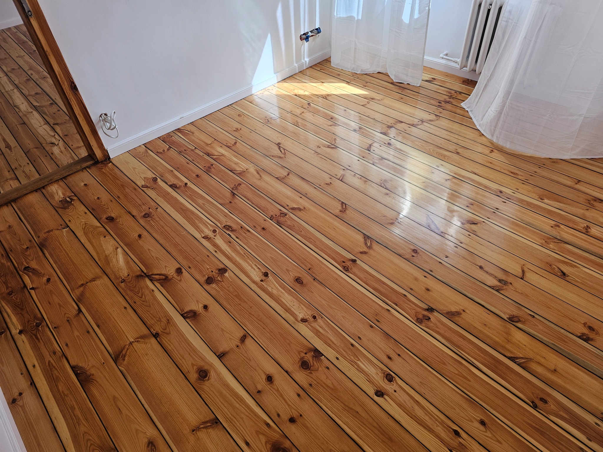 Świeżo polakierowana, jasnobrązowa podłoga z desek, ułożona ukośnie, z widocznymi sękami i naturalnym usłojeniem drewna, w rogu pokoju biała ściana z gniazdkiem elektrycznym i fragmentem okna...