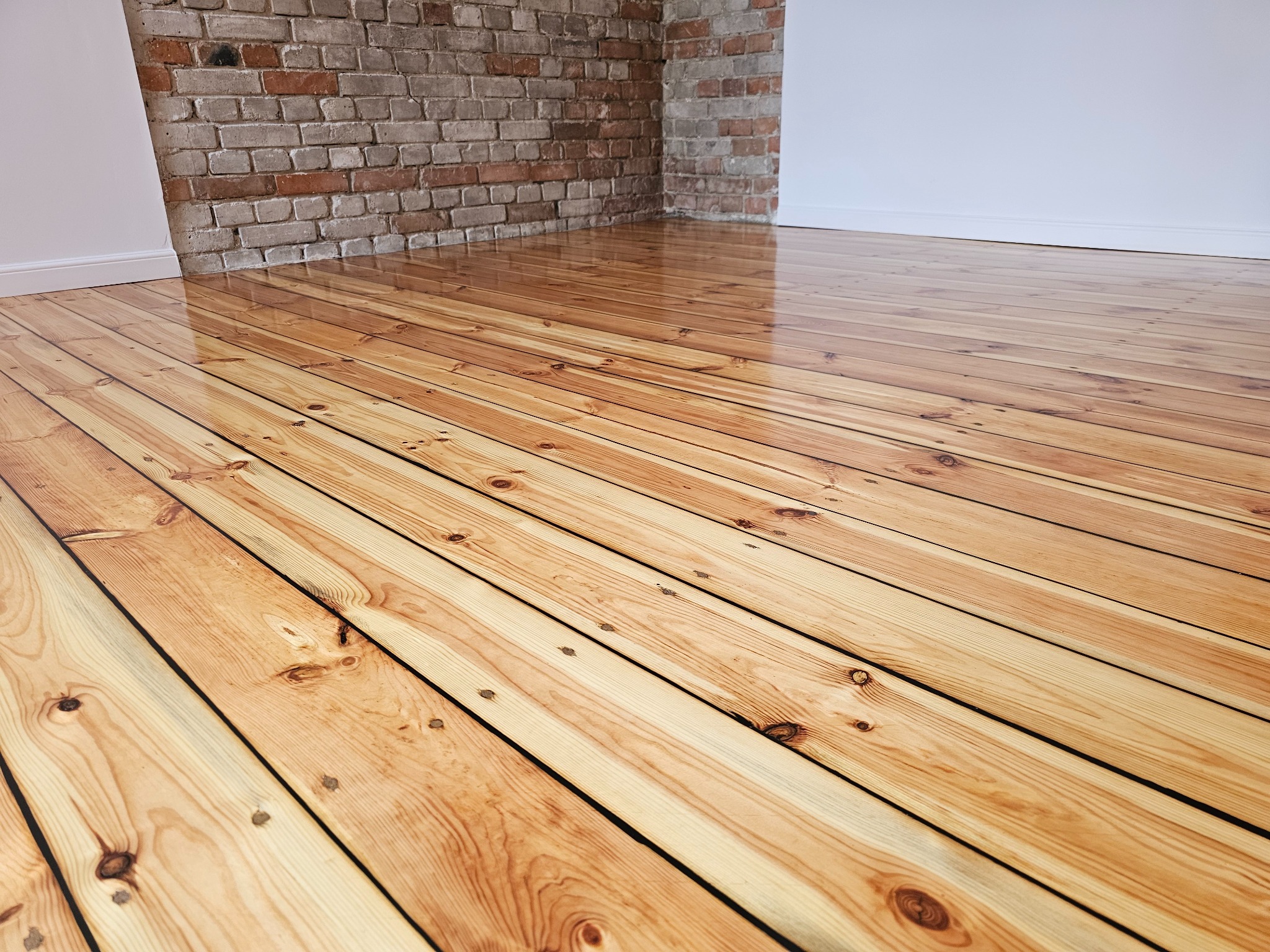 Świeżo polakierowana podłoga z desek z widocznymi słojami drewna, ceglana ściana w tle i białe listwy przypodłogowe