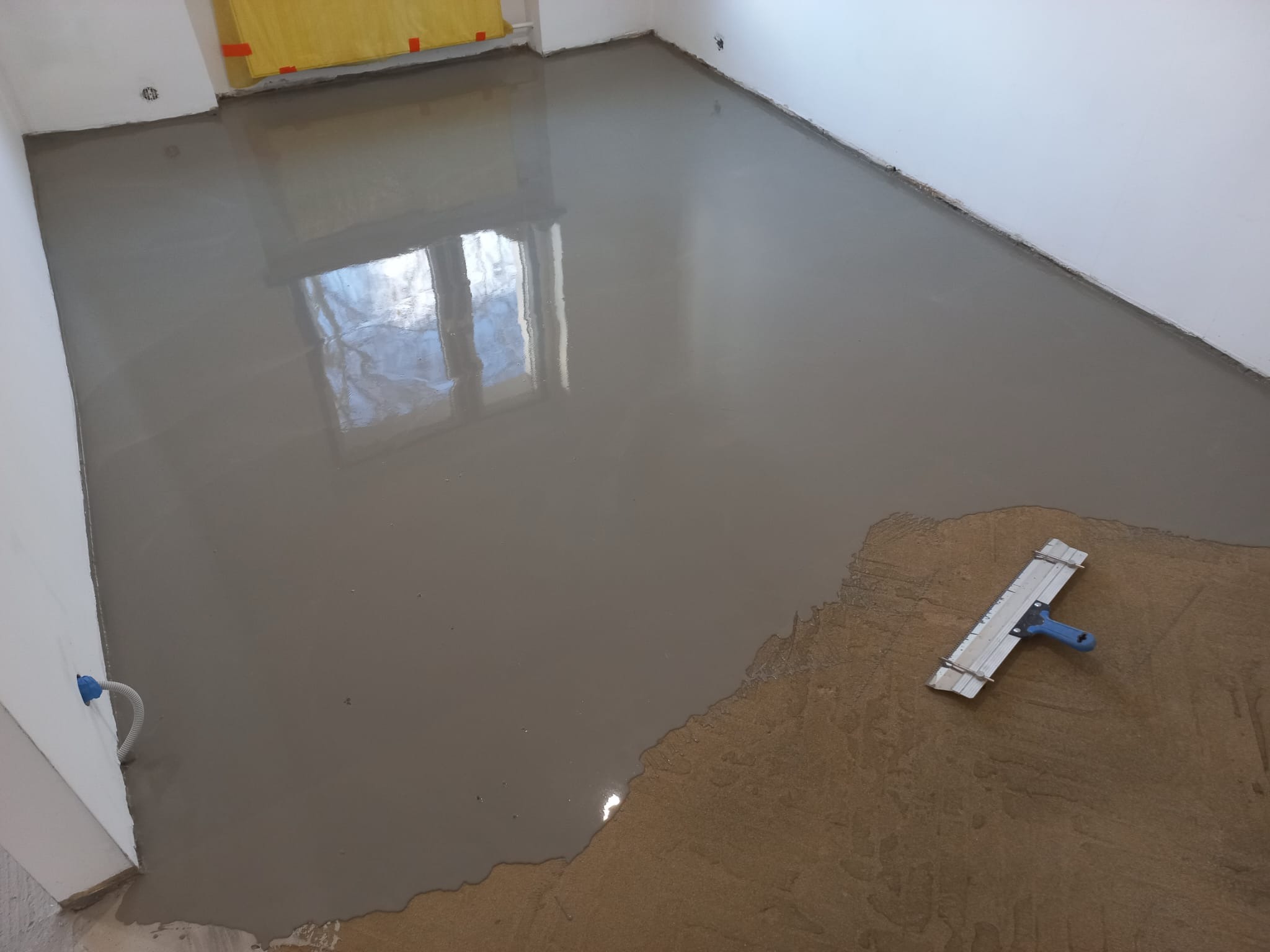 Wylewka samopoziomująca w trakcie rozprowadzania szpachlą z niebieską rączką w jasnym pomieszczeniu z oknem odbijającym się w mokrej powierzchni.