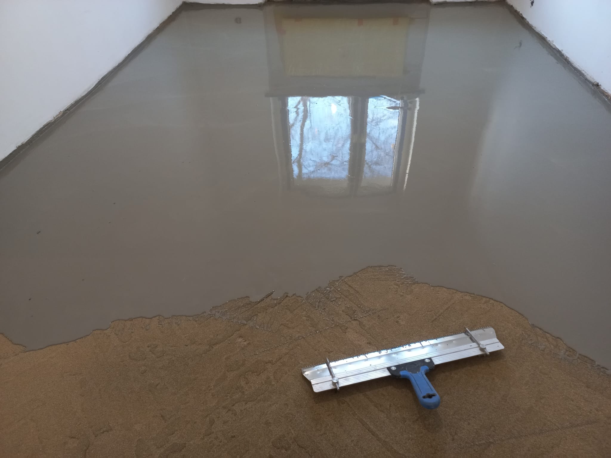 Wylewka samopoziomująca na podłodze w trakcie prac, widoczne narzędzie do rozprowadzania masy i refleksja okna w mokrej powierzchni.