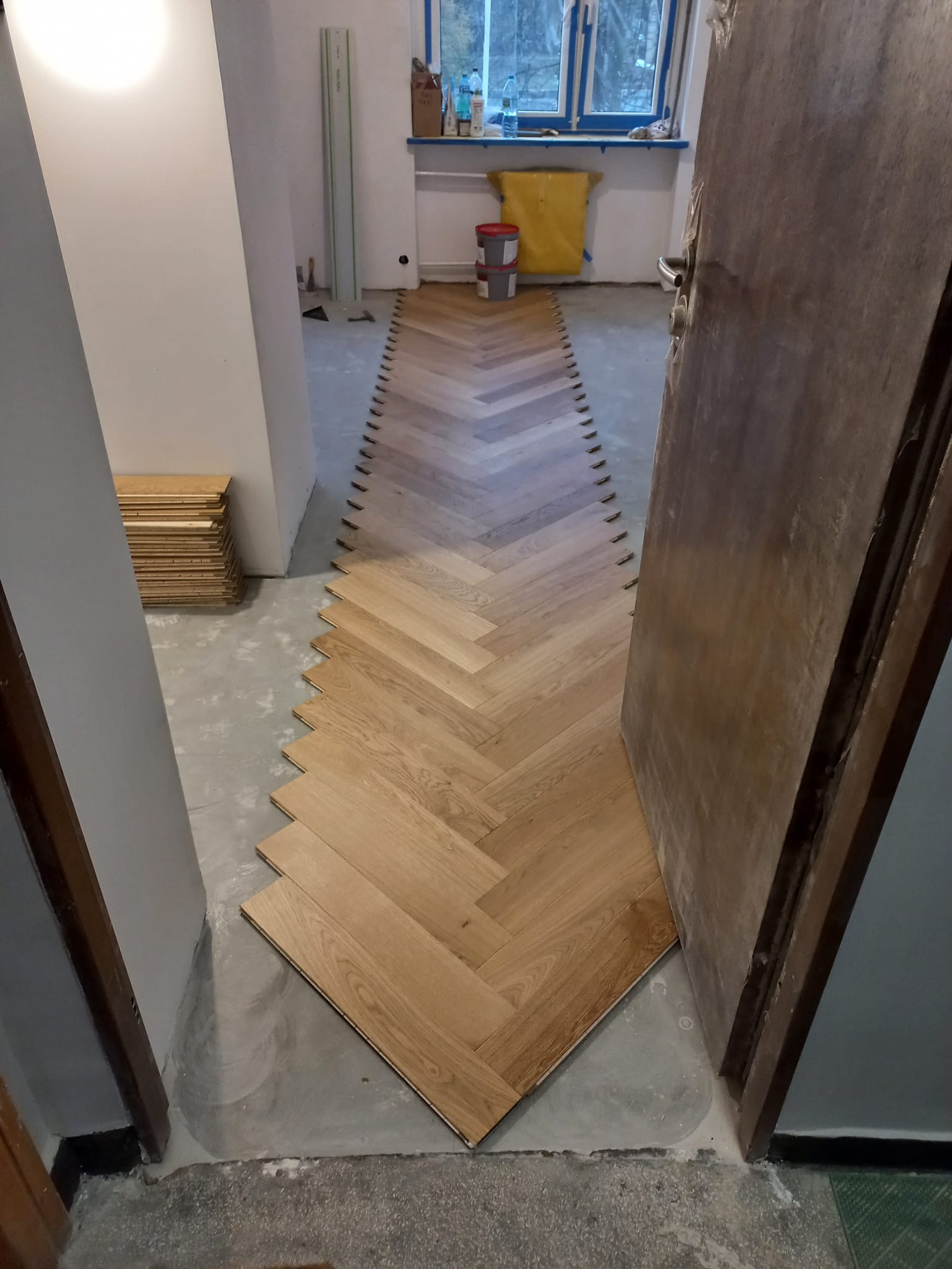 Układanie parkietu w jodełkę: widoczny postęp prac, fragment ułożonej podłogi, stos desek i listwy dystansowe, surowy beton podłoża.