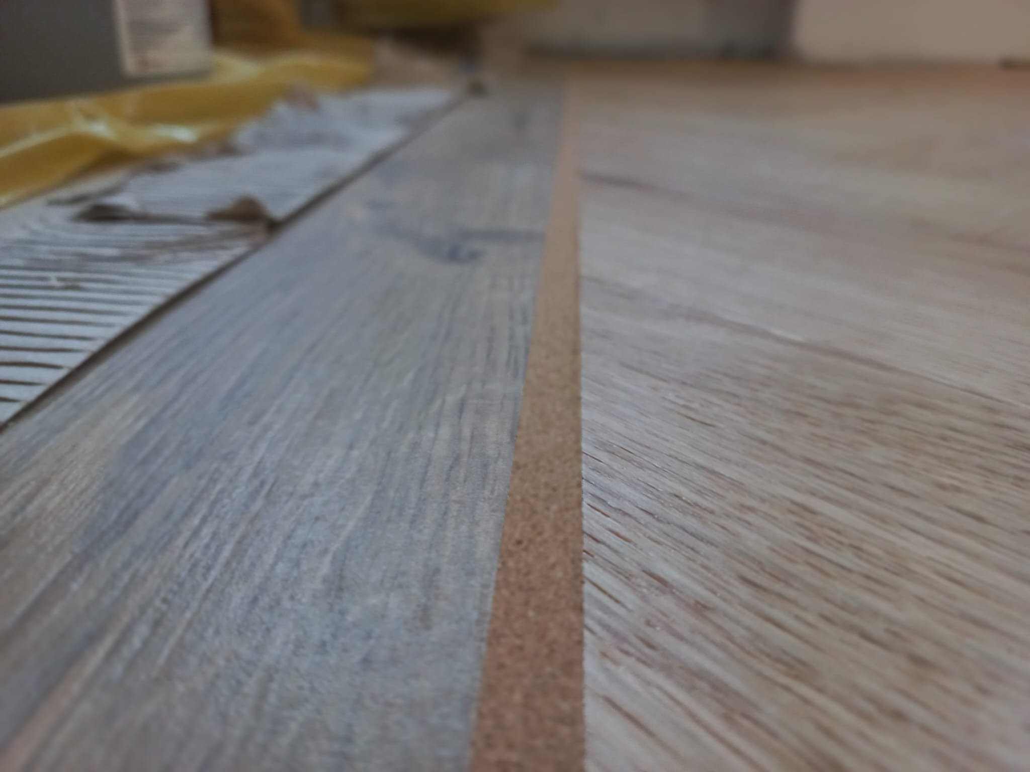 Fragment podłogi z paneli z widoczną listwą dylatacyjną i folią ochronną w tle, ujęcie z niskiej perspektywy.