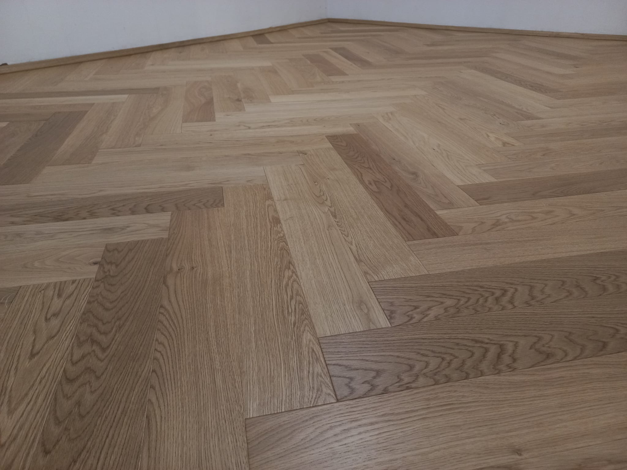 Ułożony parkiet w jodełkę w odcieniach jasnego brązu, widoczne naturalne usłojenie drewna i subtelne różnice w kolorze poszczególnych klepek, przy ścianie listwa przypodłogowa.