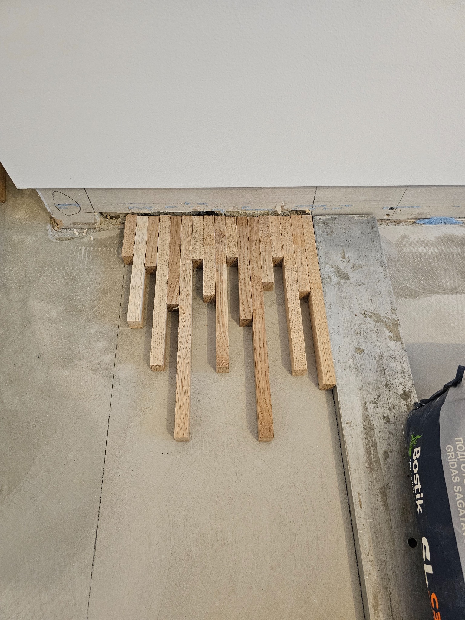 Fragment układania drewnianej podłogi z parkietu o nieregularnych długościach, widoczny worek z klejem Bostik, surowy betonowy podkład.