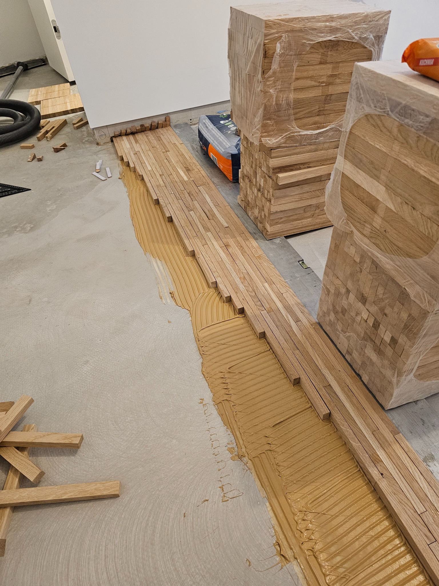 Fragment podłogi z klejem i ułożonymi, jasnymi, drewnianymi klepkami parkietu; w tle stosy zapakowanych elementów oraz rura od odkurzacza.