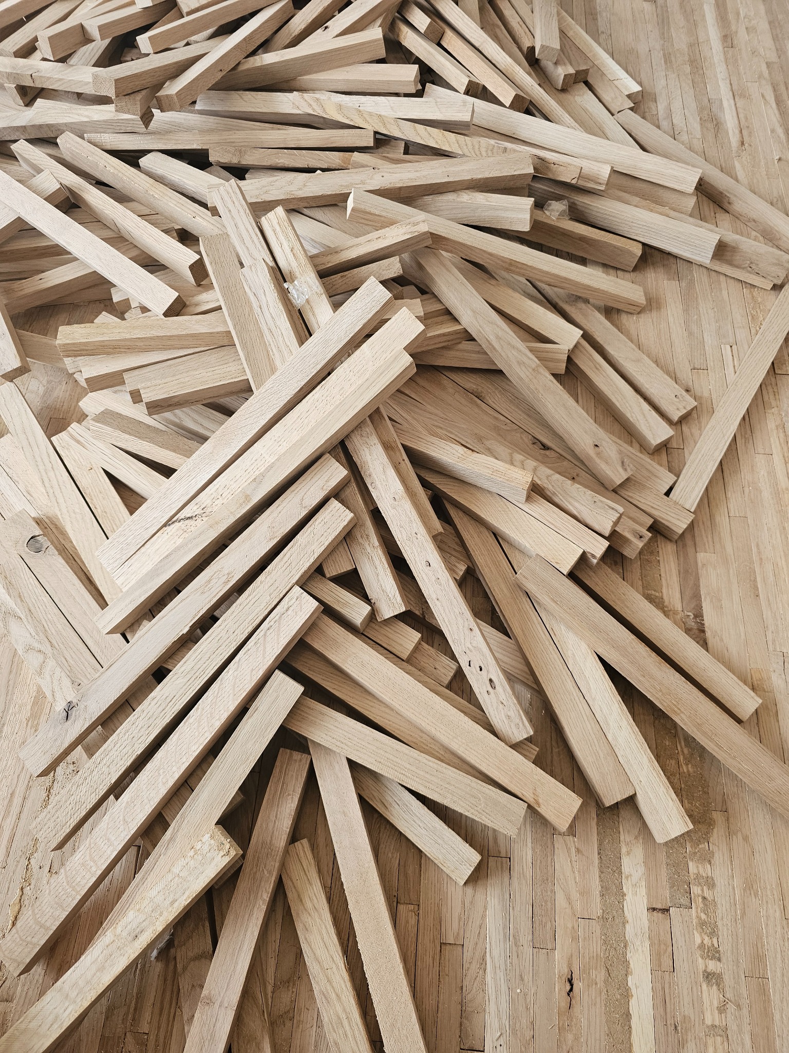 Stos surowych, jasnych listew parkietowych z widocznymi słojami drewna, rozrzuconych na istniejącej, częściowo uszkodzonej drewnianej podłodze.