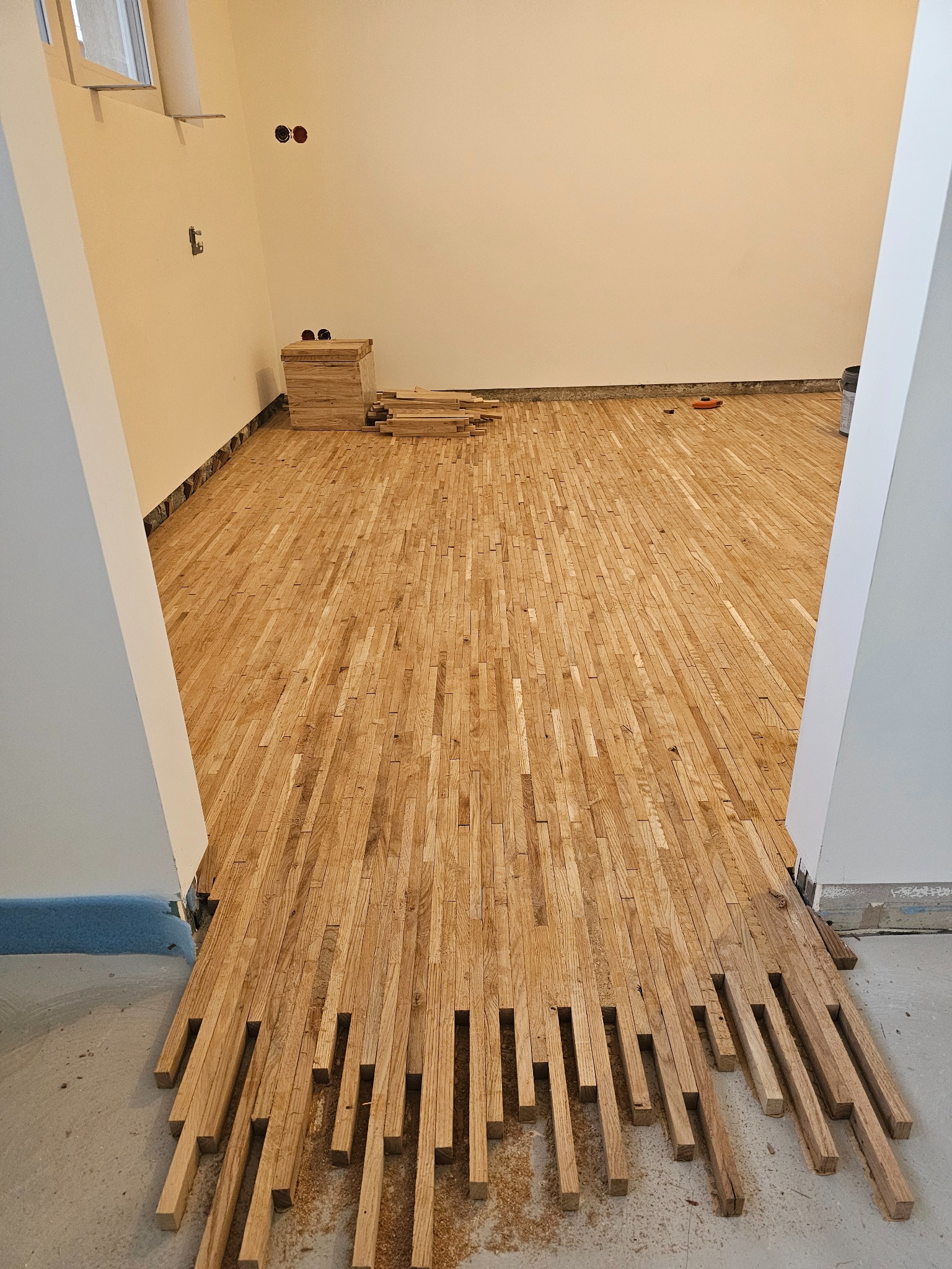 Układanie parkietu z krótkich, prostokątnych elementów w surowym stanie, widoczne niedokończone fragmenty przy ścianach i stosy nieułożonych elementów.