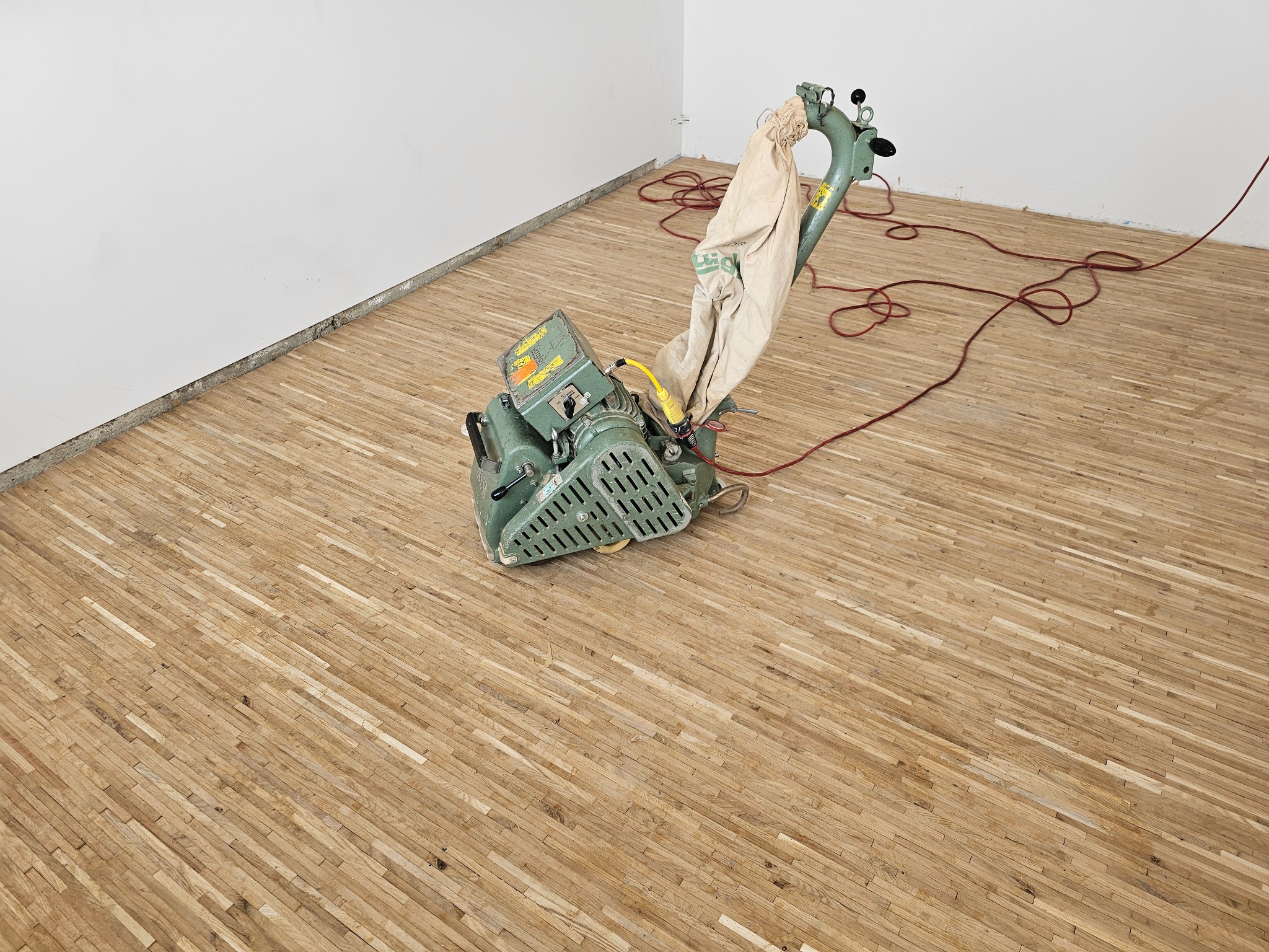 Szlifowanie drewnianej podłogi parkietowej za pomocą cykliniarki, widoczny worek na pył i przewód zasilający.