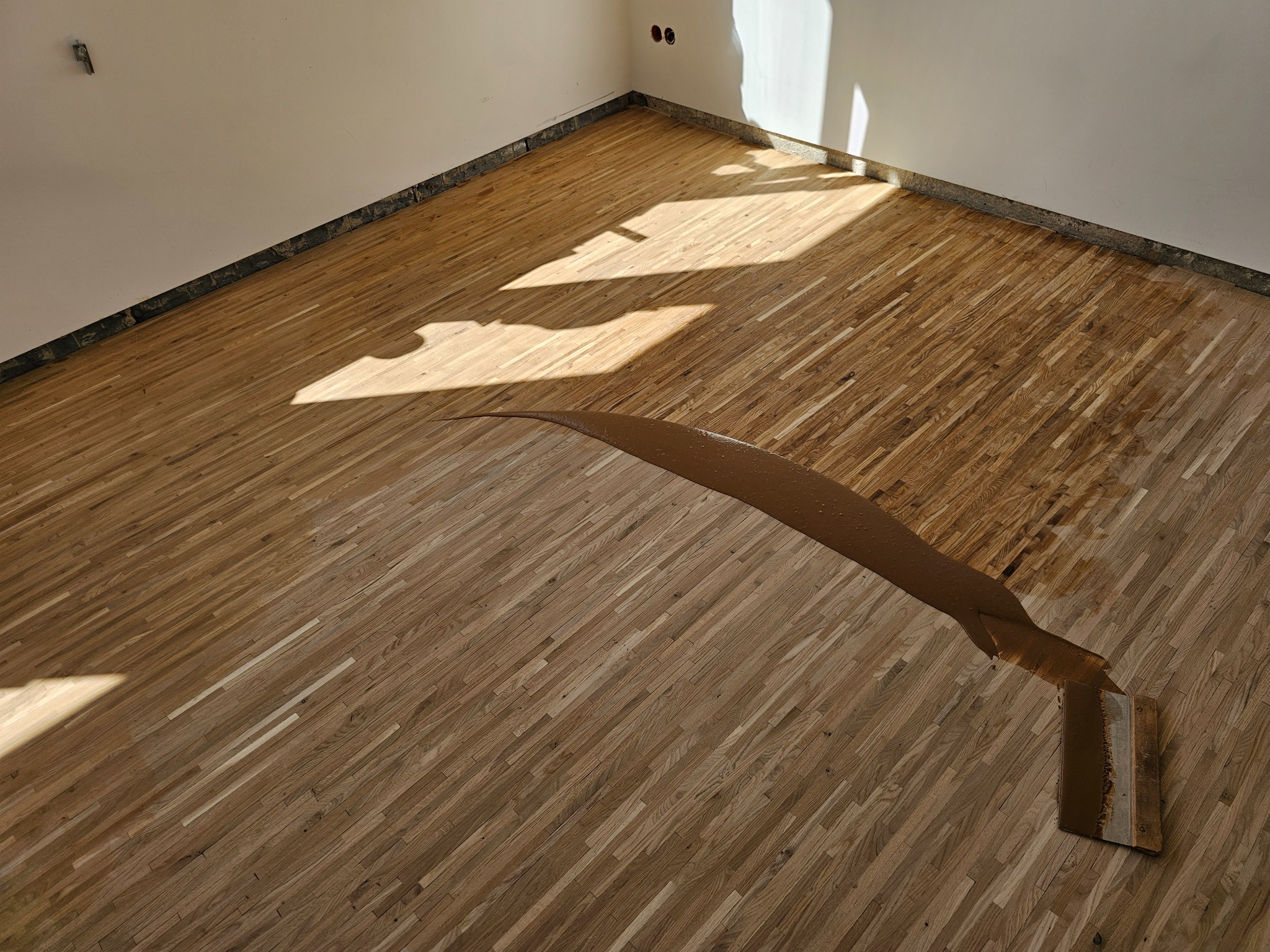 Aplikacja brązowego lakieru na drewniany parkiet w rogu pokoju, widoczna szpachla i świeże wykończenie podłogi w jasnym świetle.