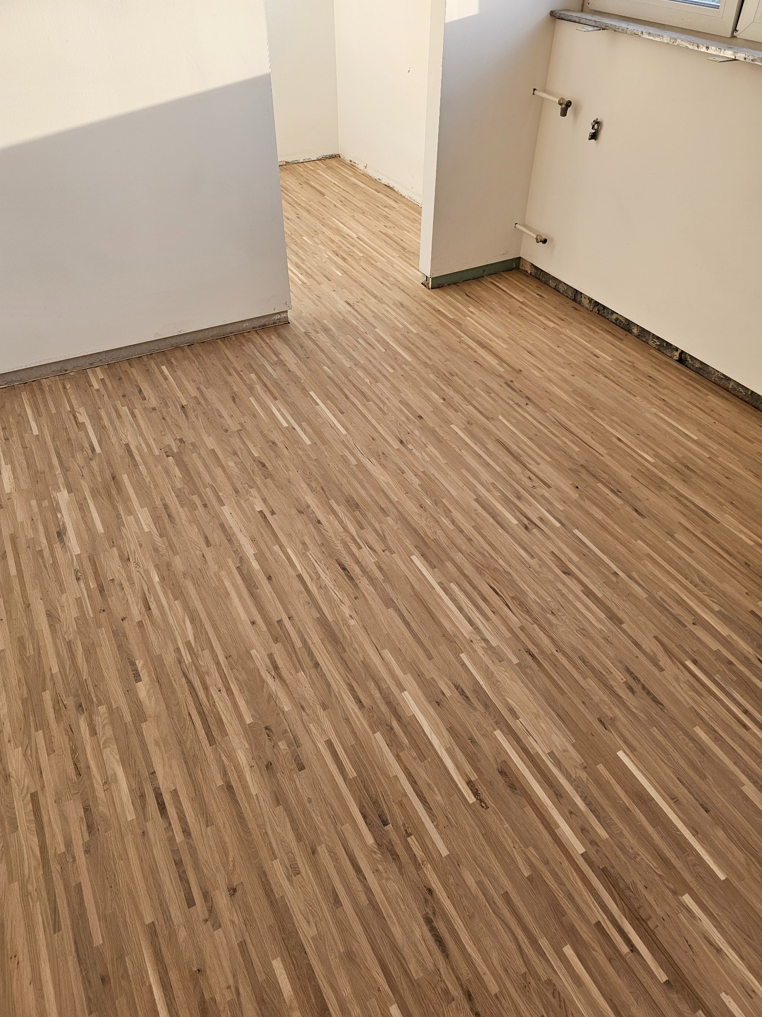 Nowo ułożona, jasnobrązowa podłoga parkietowa w surowym wnętrzu. Widoczne niedokończone ściany i wystające rury.