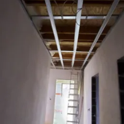 Wnętrze w trakcie montażu metalowej konstrukcji pod sufitem podwieszanym, widoczna drabina i przewody elektryczne.