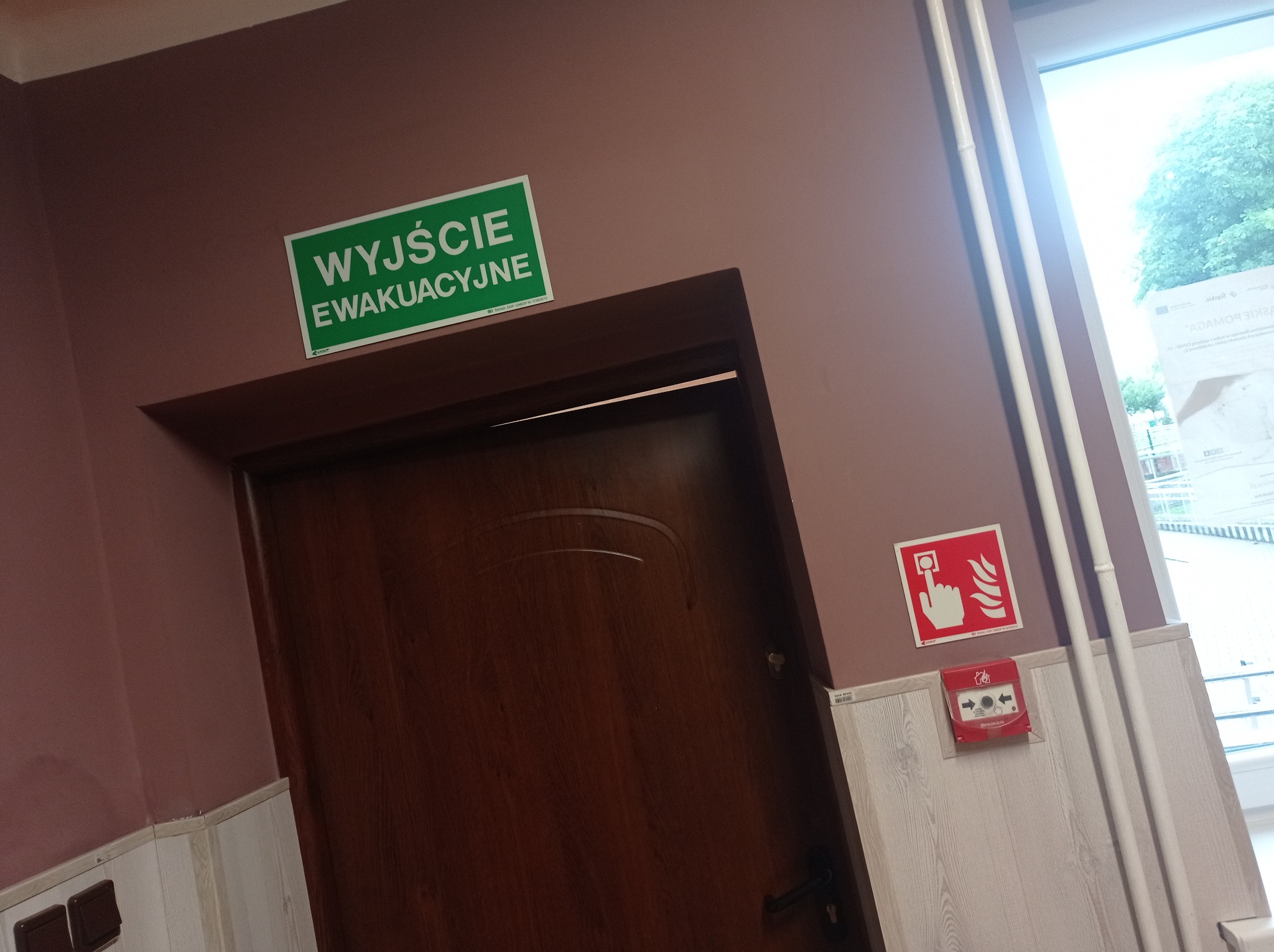 Brązowe drzwi z zielonym znakiem 'Wyjście Ewakuacyjne' nad nimi, czerwony przycisk alarmowy na ścianie z boazerią, widoczne rury grzewcze i okno w tle.