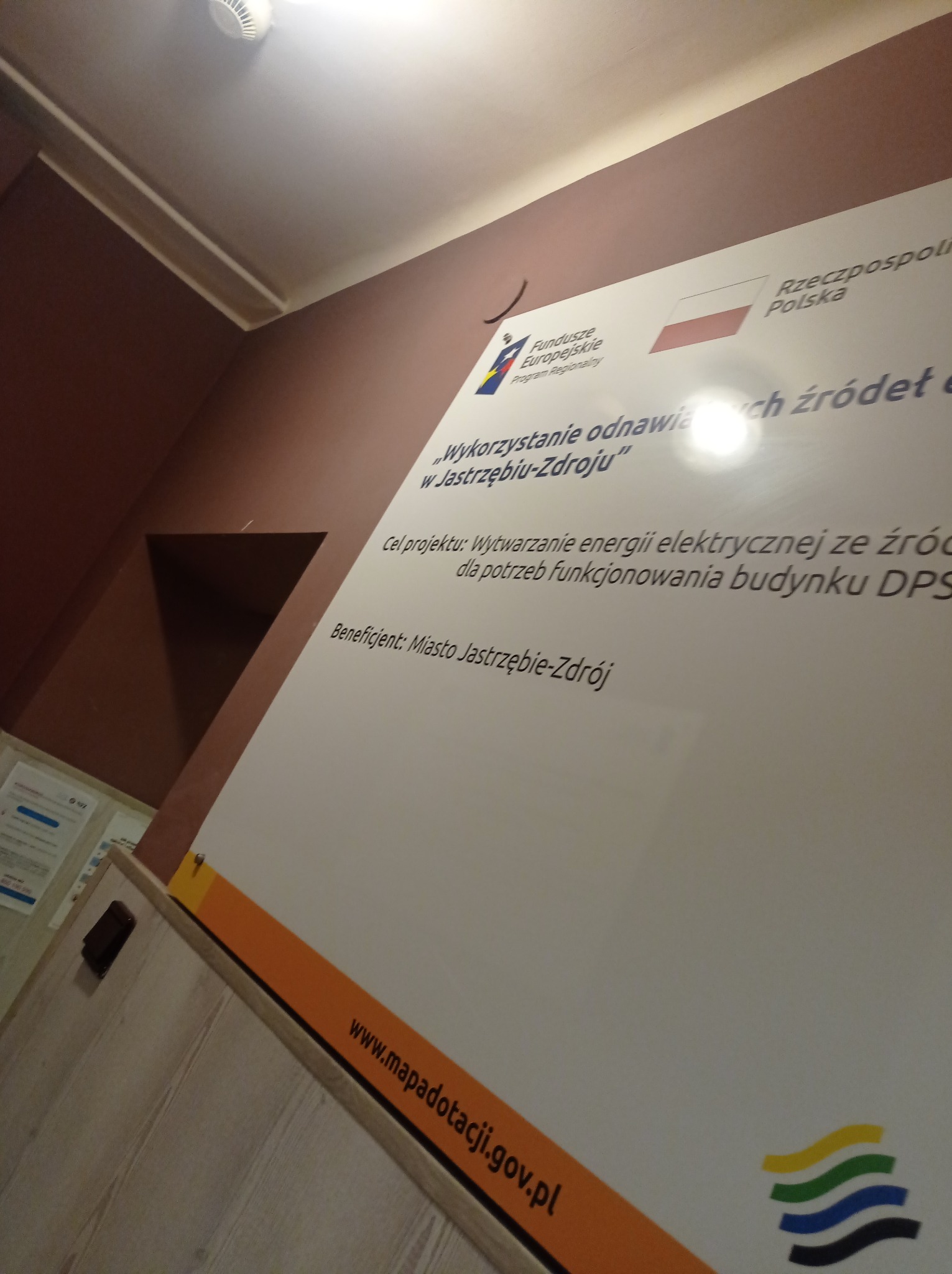 Tablica informacyjna na ścianie, prezentująca projekt 'Wykorzystanie odnawialnych źródeł energii w Jastrzębiu-Zdroju', z logo Funduszy Europejskich, flagą Polski i adresem strony internetowej www.