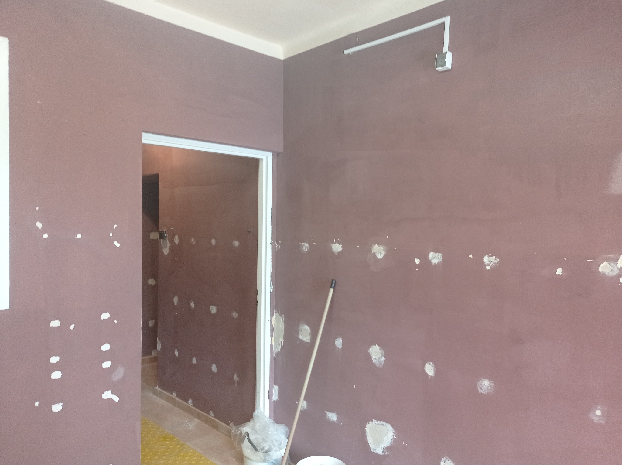 Przygotowanie ściany do malowania: szpachlowanie ubytków w fioletowej ścianie, widoczny wałek malarski i wiadro z gładzią.