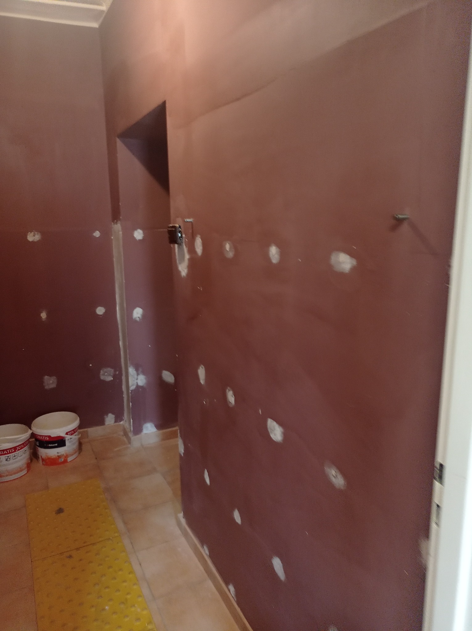 Ściana w trakcie szpachlowania z widocznymi łatami gładzi, przygotowywana do malowania, z dwoma wiadrami farby w rogu pomieszczenia i żółtą matą ochronną na podłodze.