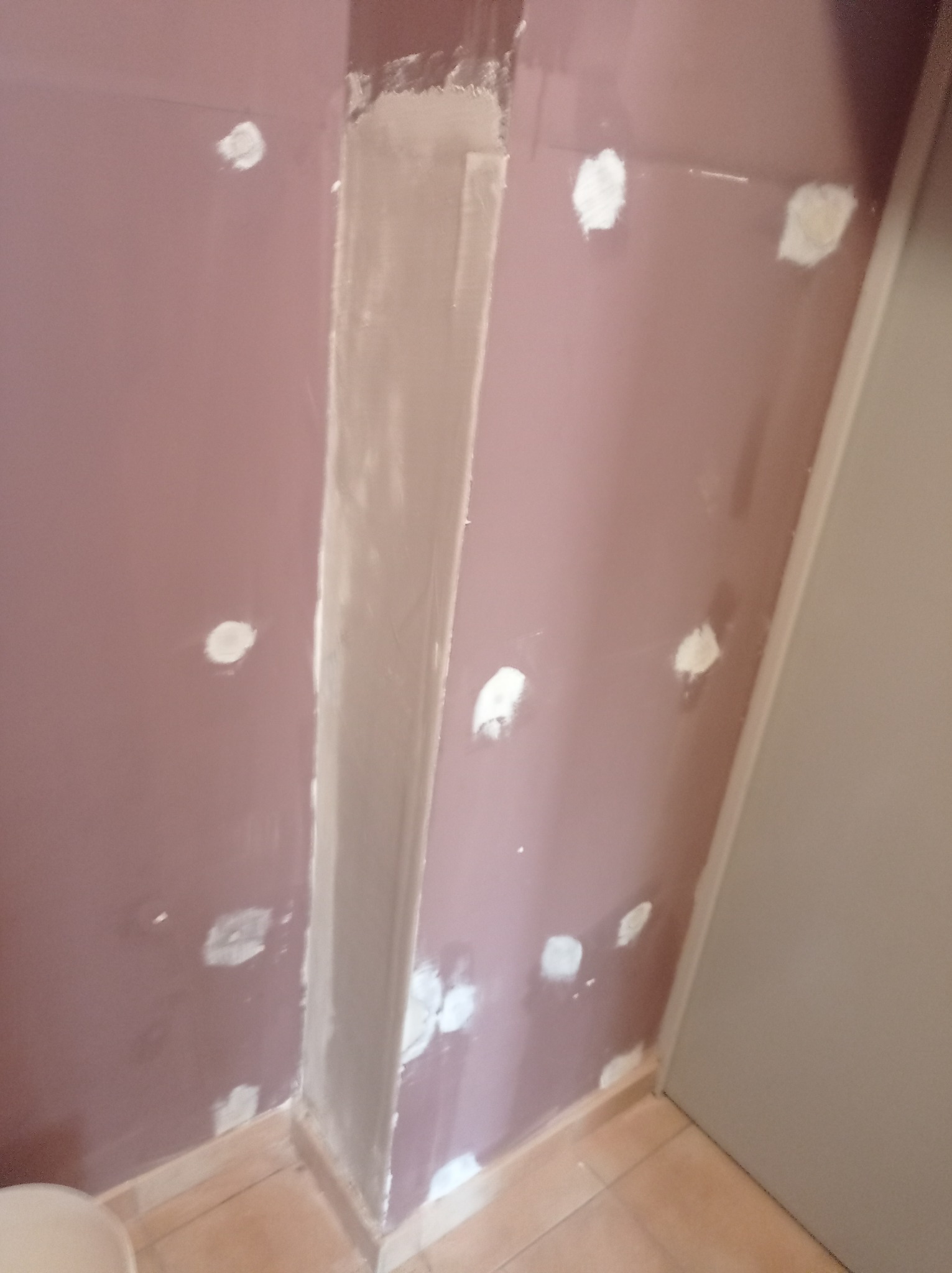 Fioletowa ściana z widocznymi punktowymi naprawami gipsowymi i świeżo wykończonym narożnikiem, płytki podłogowe w dolnej części kadru.