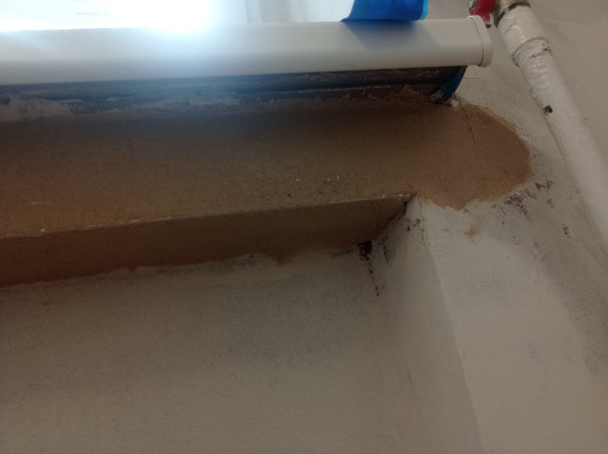 Przygotowanie do malowania okna: zaklejona taśmą rama okienna, widoczne ślady szpachlowania i rura instalacyjna przy ścianie.