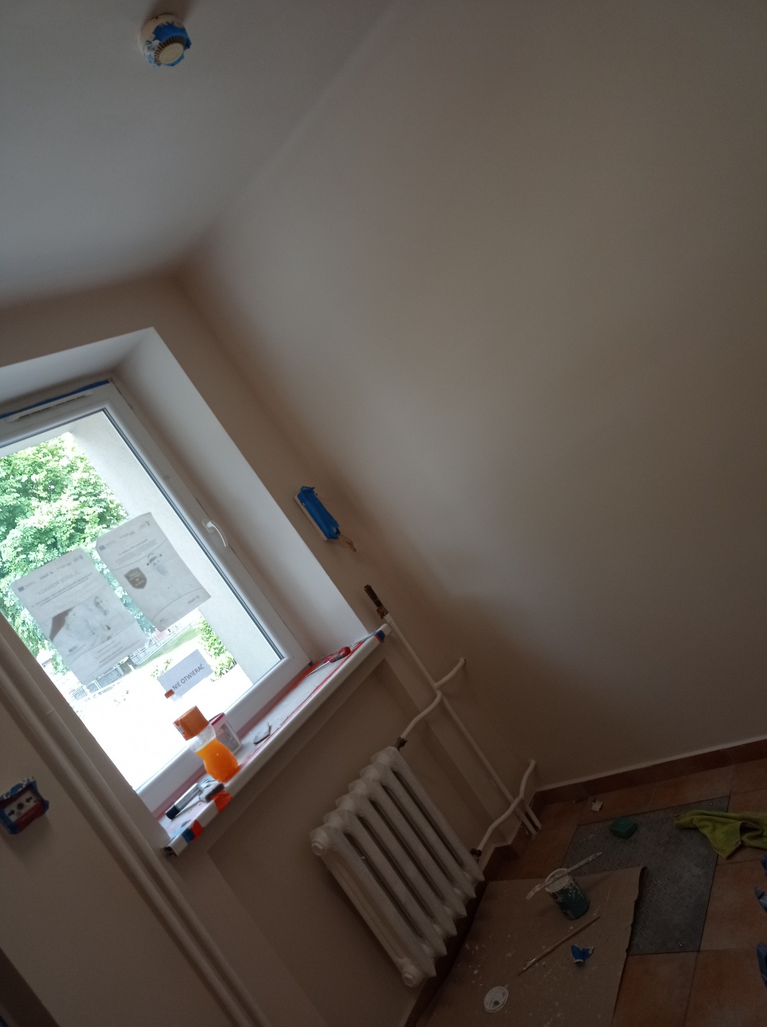 Świeżo pomalowane ściany w pokoju z zabezpieczonym czujnikiem dymu, widoczne narzędzia malarskie, grzejnik i okno z naklejonymi kartkami.
