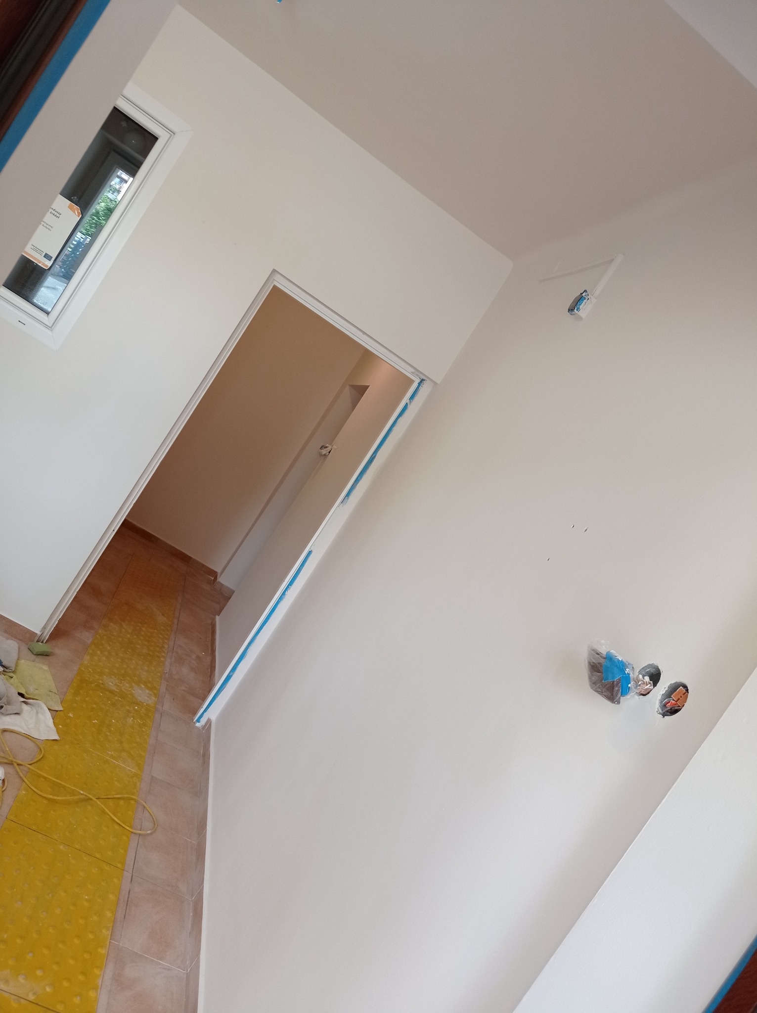 Świeżo pomalowane na biało ściany w pomieszczeniu z widocznymi zabezpieczeniami taśmą malarską wokół okna i drzwi, żółta folia ochronna na podłodze oraz wystające kable elektryczne.