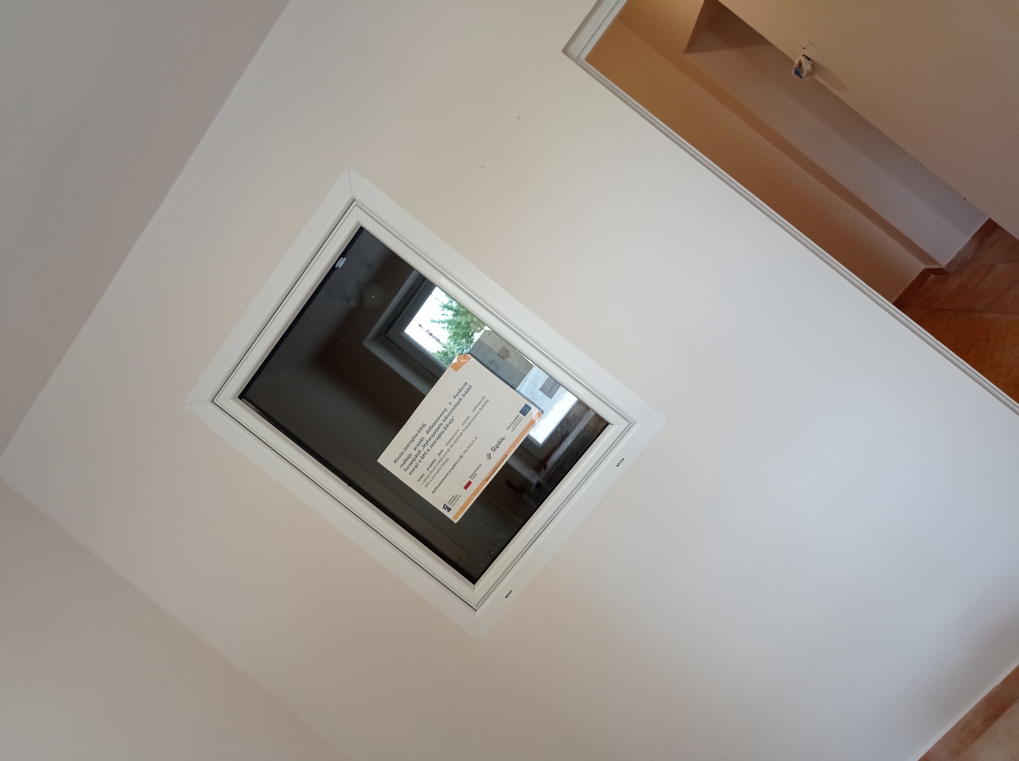 Okno dachowe z naklejoną informacją o dofinansowaniu projektu z funduszy europejskich w jasnym wnętrzu. Widoczny fragment pomieszczenia obok z drewnianą podłogą.