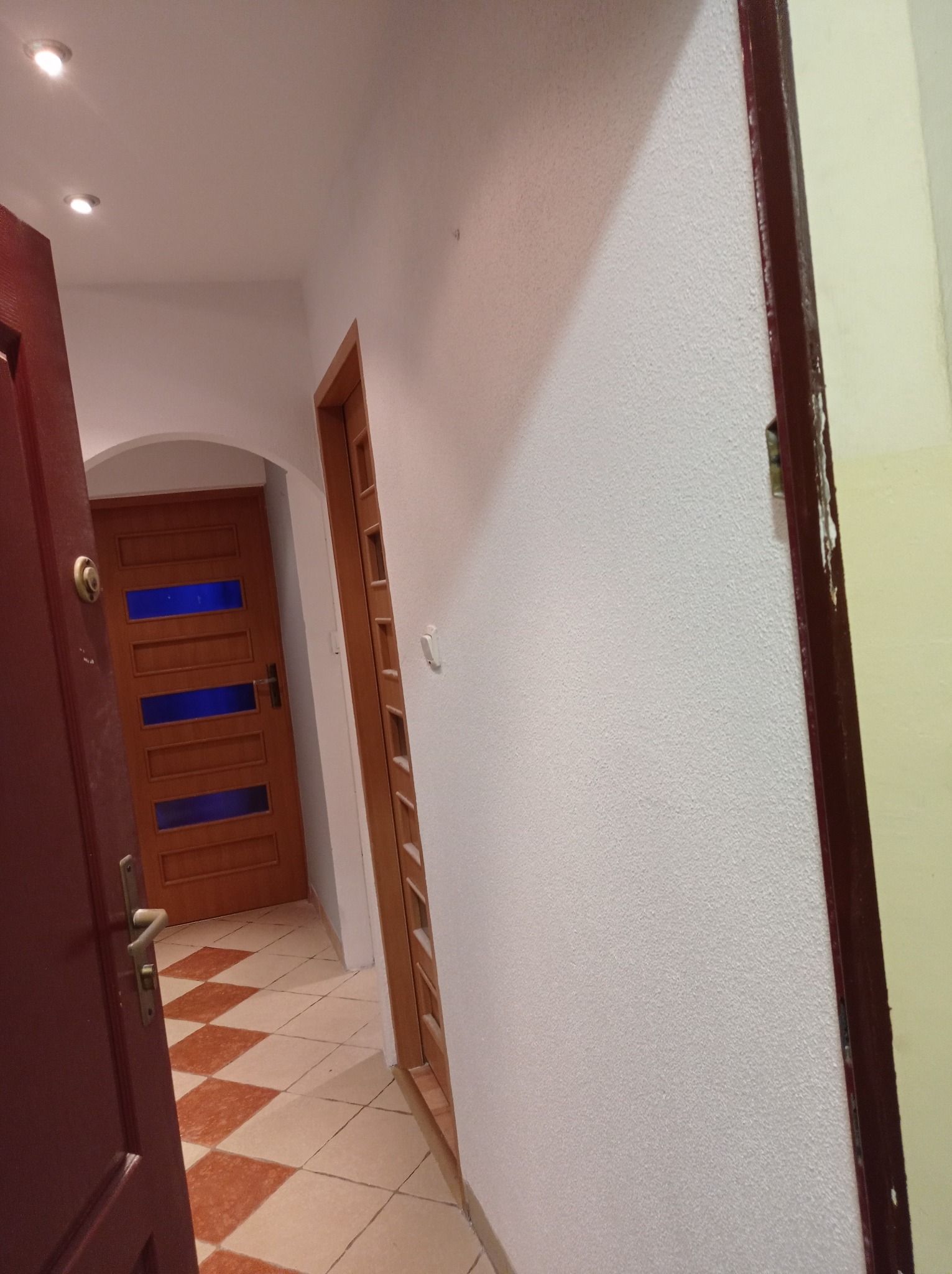 Korytarz po odświeżeniu: pomalowane ściany na biało z widoczną fakturą, terakota w brązowo-białą szachownicę, dwie drewniane ościeżnice i częściowo widoczna, otwarta, ciemnoczerwona drewniana brama...