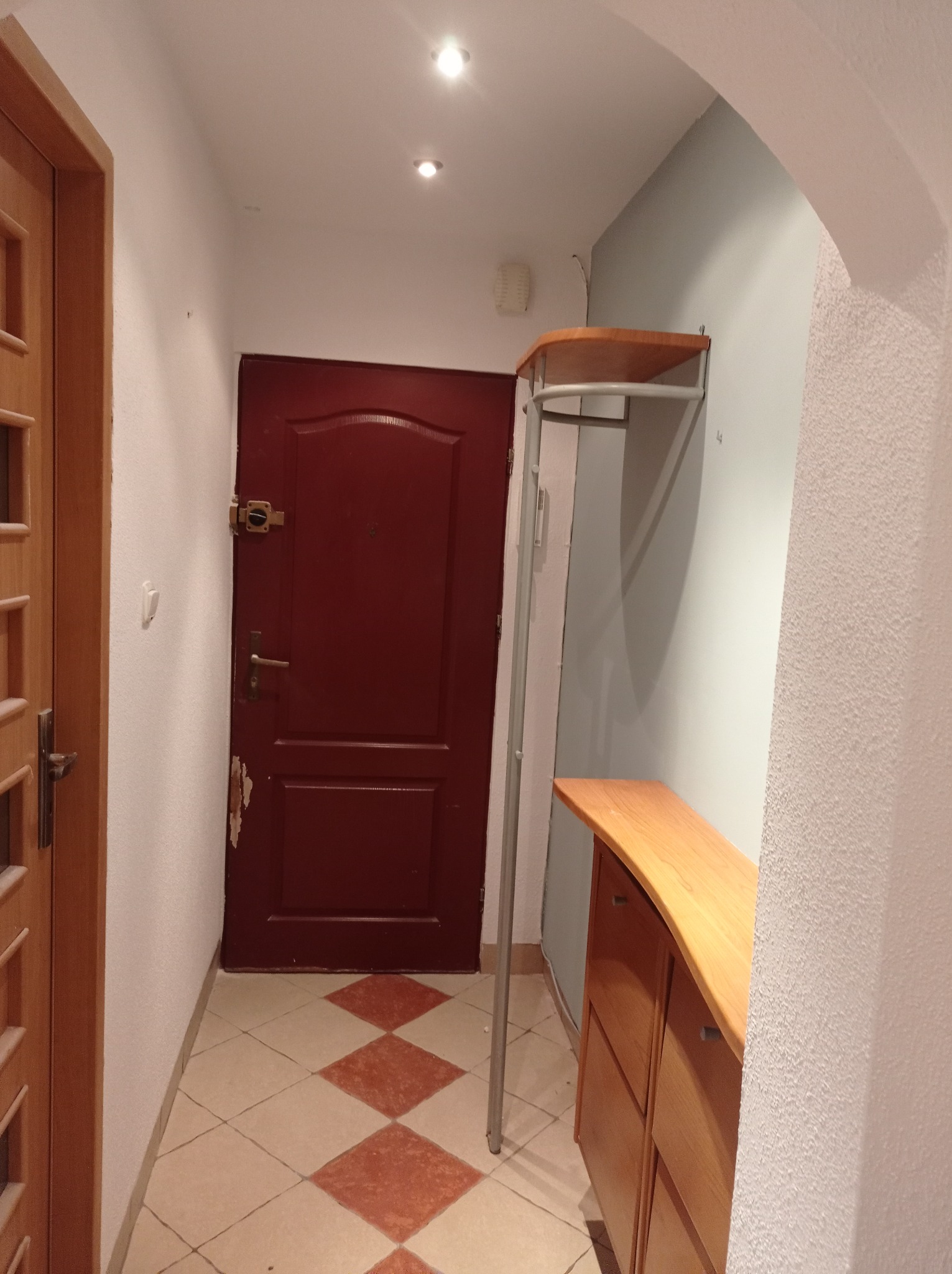 Przedpokój z brązowymi drzwiami wejściowymi, szafką na buty z jasnego drewna oraz podłogą w beżowo-brązową szachownicę. Ściany białe i szare, widoczne ślady uszkodzeń przy drzwiach.