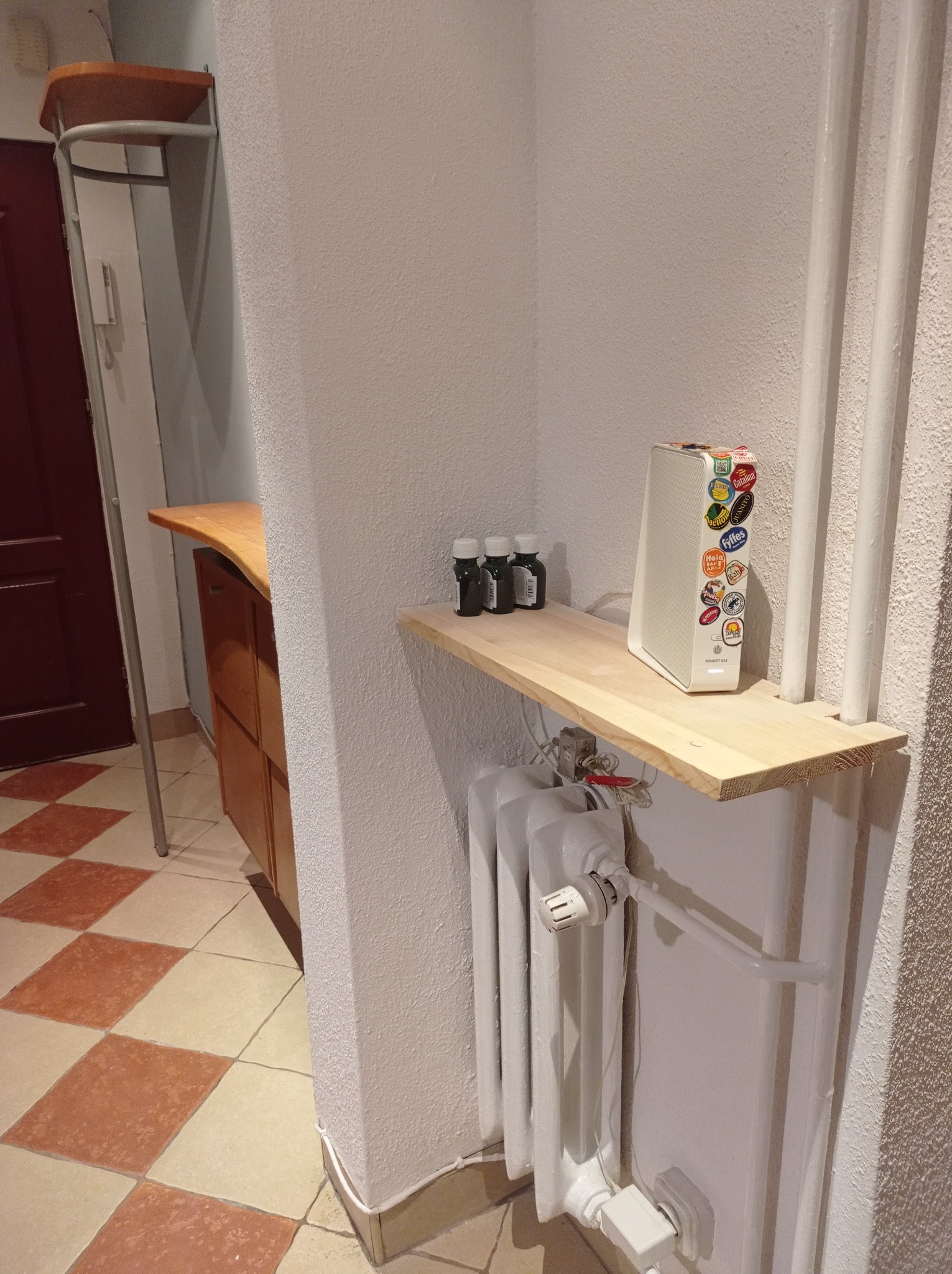 Półka z surowego drewna zamontowana nad grzejnikiem w przedpokoju, na niej trzy małe butelki i biały router obklejony naklejkami.