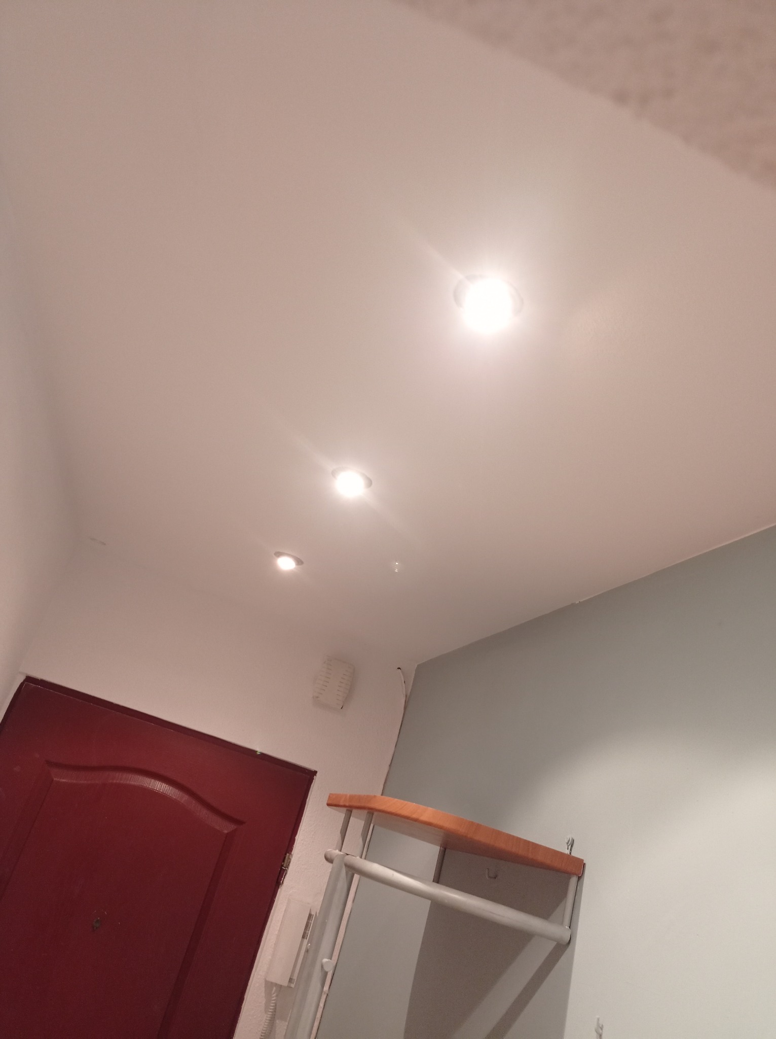 Wnętrze przedpokoju z trzema punktowymi lampami sufitowymi, częściowo widoczna czerwona drzwi i fragment ściany z wieszakiem.