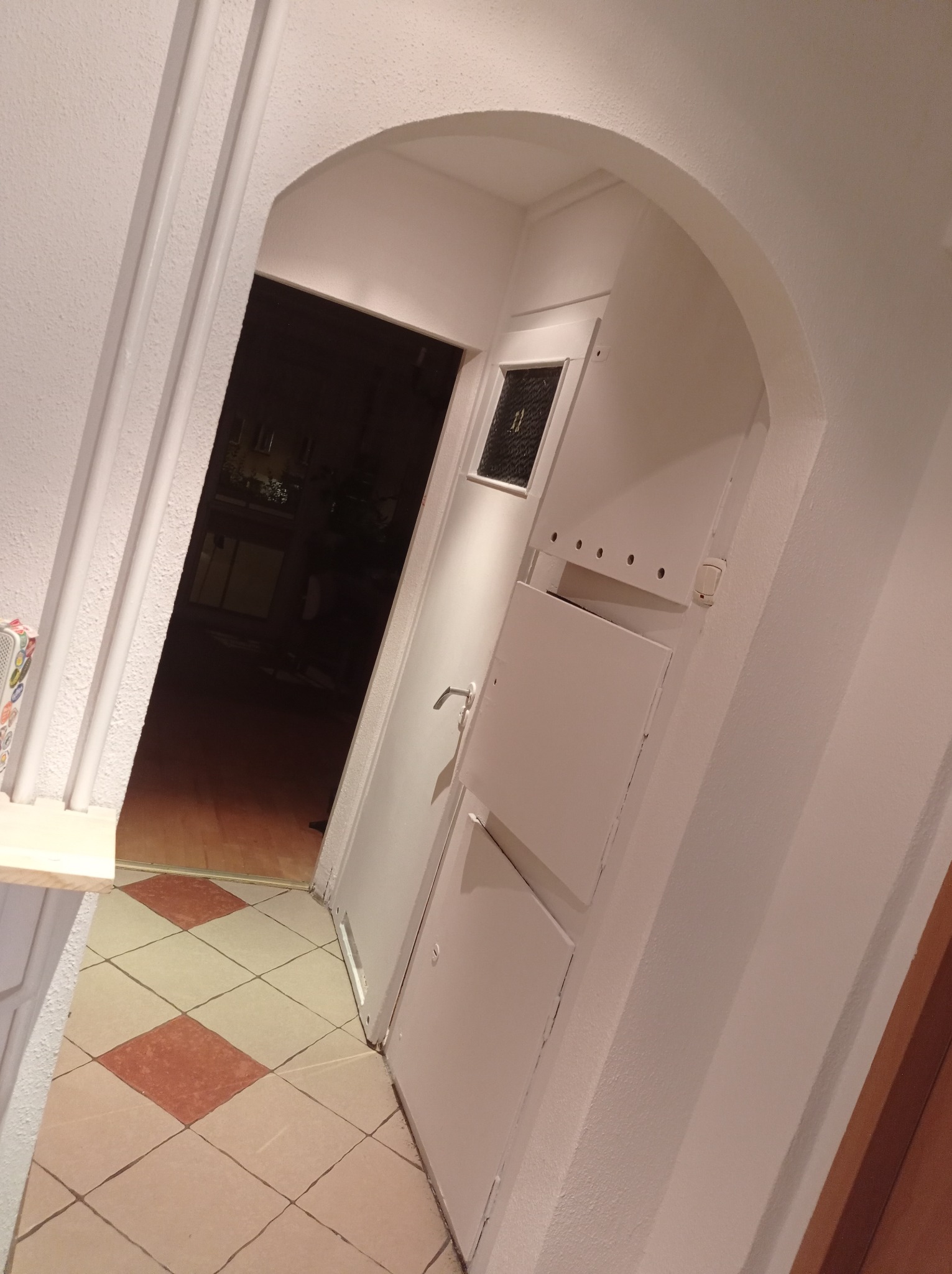 Wejście do mieszkania z łukowatym przejściem, białe ściany, drzwi z szybką i panelami, podłoga z kwadratowych płytek w kolorze beżowym i brązowym.