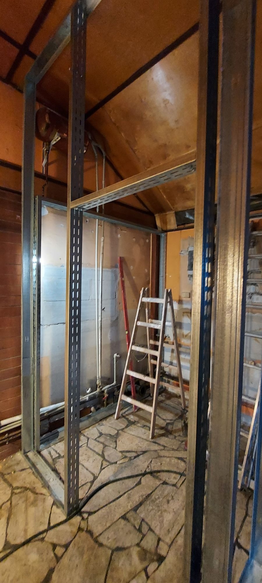 Metalowa konstrukcja szkieletowa w trakcie montażu, widoczna drabina i fragmenty instalacji, podłoga wyłożona kamiennymi płytami o nieregularnych kształtach.