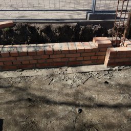 Budowa Marzeń - Wznoszenie muru z czerwonej cegły na fundamencie z widocznym zbrojeniem, wykop pod fundament w tle.