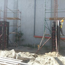 Budowa Marzeń - Prace ziemne pod budowę, szalunki kolumnowe zbrojone stalą, ustawione na placu budowy, pracownik w kasku i kamizelce na rusztowaniu, w tle szara ściana budynku.