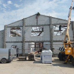 Budowa Marzeń - Budowa budynku z białych bloczków, widoczne rusztowania, dźwig, samochód dostawczy i palety z materiałem budowlanym firmy Prefabet, na szczycie budynku wieniec.