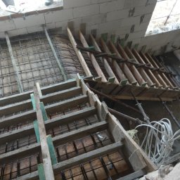 Budowa Marzeń - Wykonywanie szalunku i zbrojenia pod fundamenty wewnątrz budynku z białych bloczków, widoczne drewniane stemple i stalowe pręty zbrojeniowe, kabel elektryczny w tle.