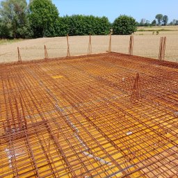 Budowa Marzeń - Zbrojenie płyty fundamentowej na deskowaniu z widocznymi wystającymi prętami, w tle pole uprawne i drzewa pod błękitnym niebem.