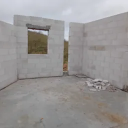 Wnętrze w stanie surowym otwartym z częściowo wzniesionymi ścianami z bloczków, widoczne okno z nadprożem i wystającymi prętami zbrojeniowymi z fundamentu, na posadzce resztki materiałów...